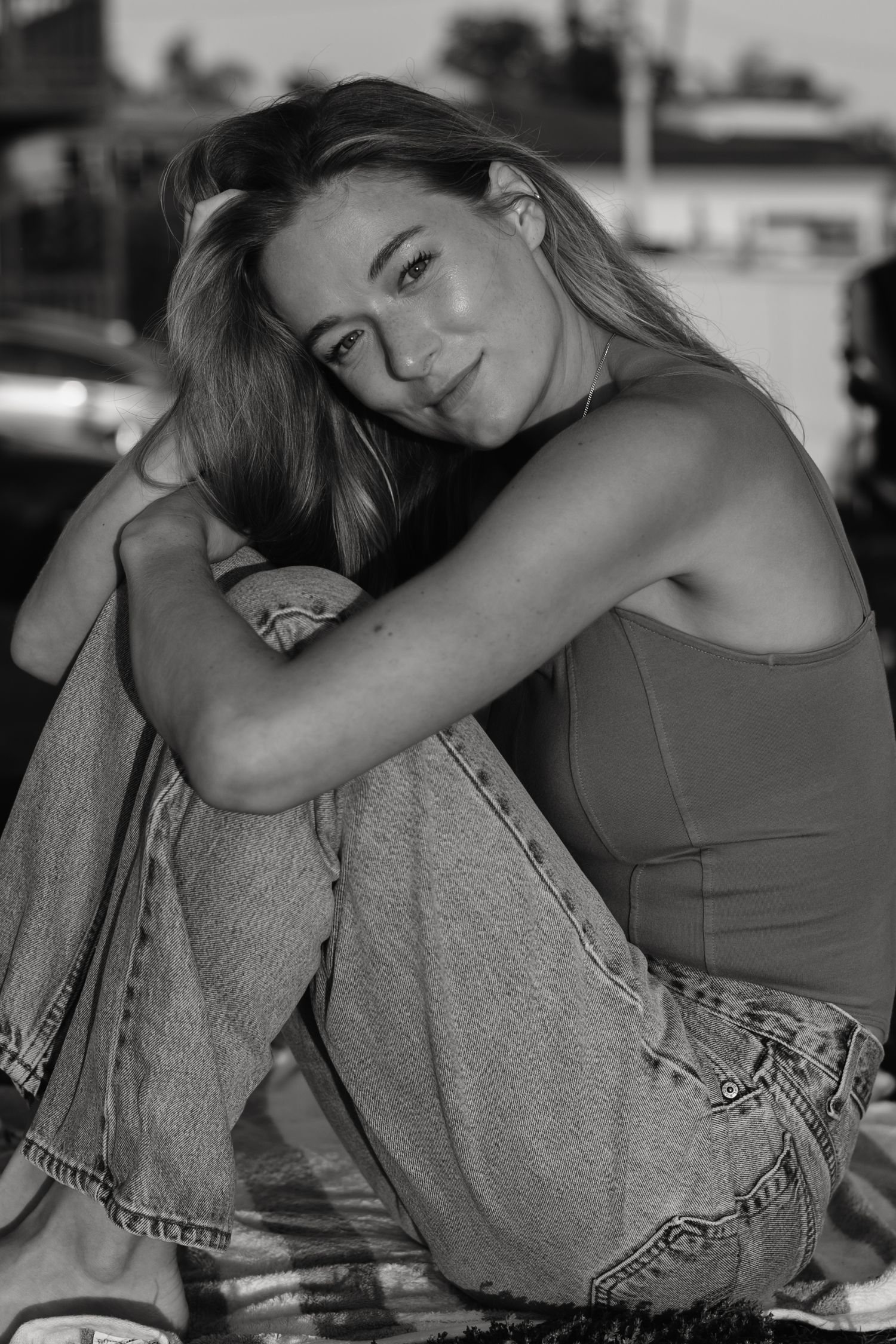 Black and white photo of a young blonde woman sitting with knees up, wearing jeans and a tank top, smiling softly.