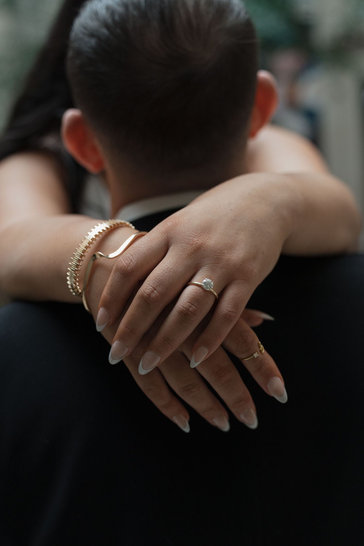 Close-up of brides hands with engagement ring and gold bracelet wrapped around grooms shoulders.