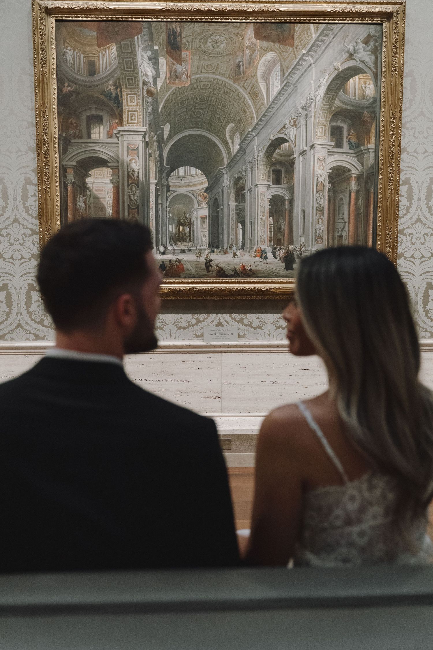 Couple in formal attire admiring a classical architectural painting in an elegant art gallery.