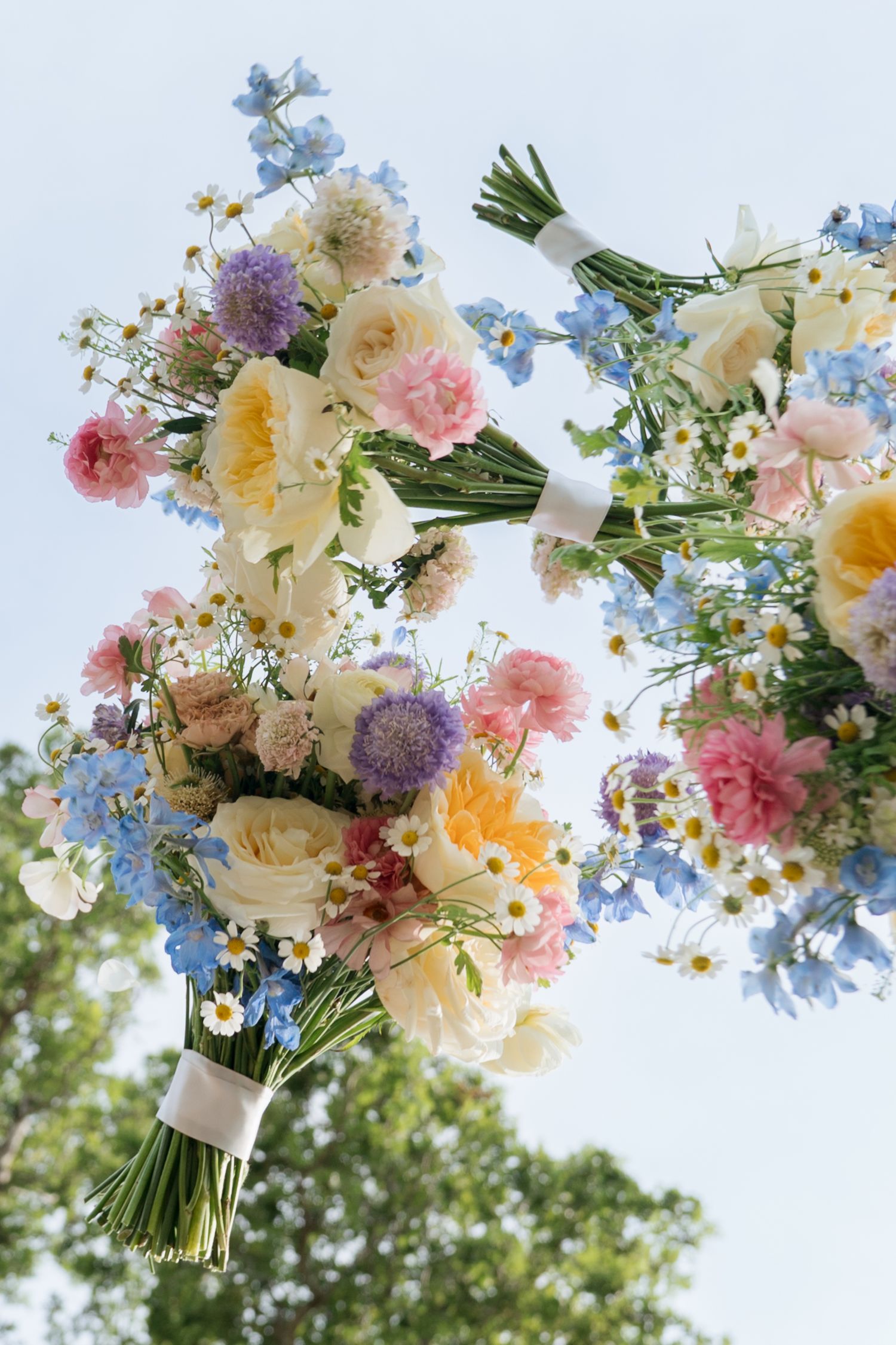 Colorful wedding bouquets with roses, daisies, and blue flowers held against a bright sky.