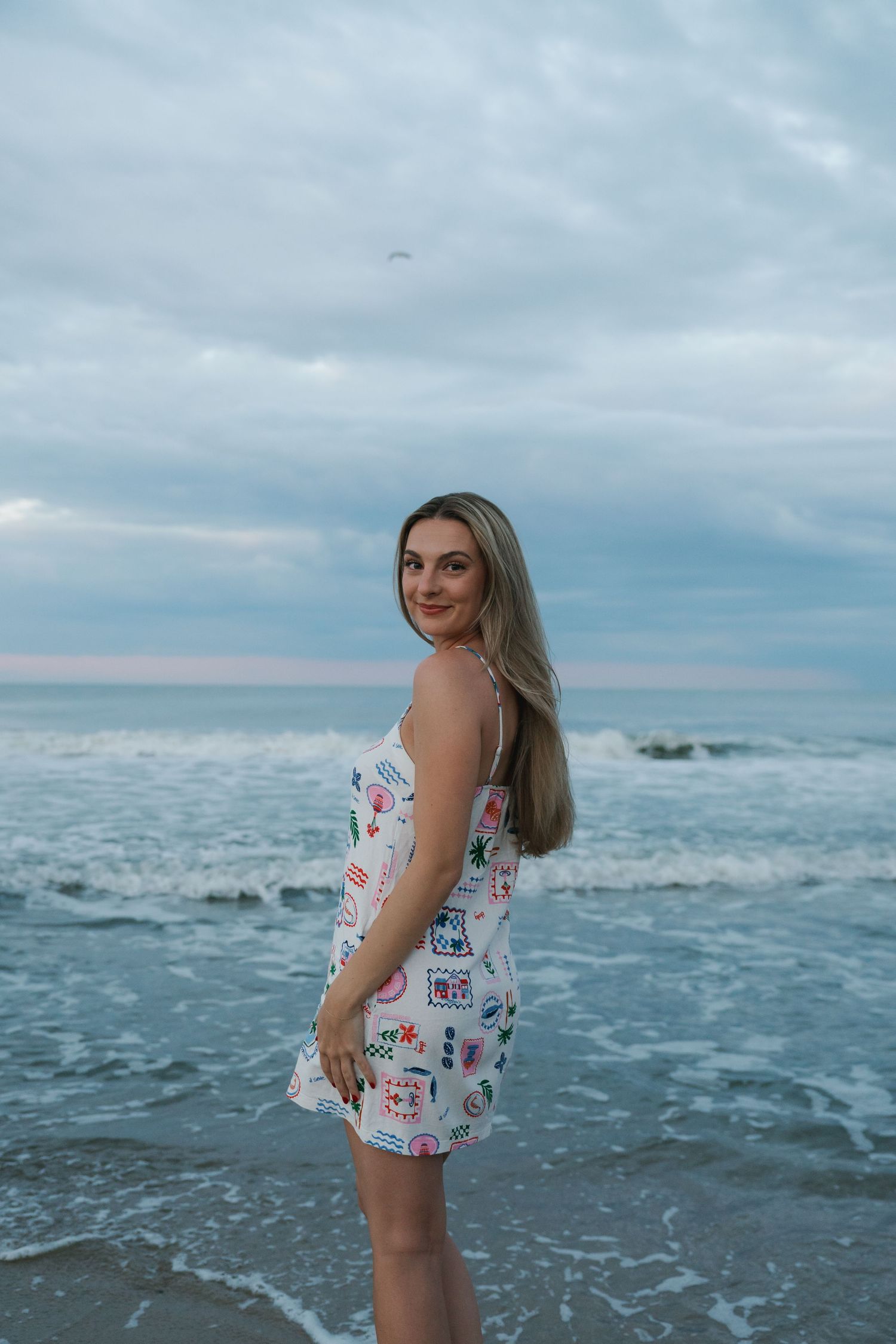 Woman in colorful printed sundress standing at ocean shore during cloudy sunset, smiling over shoulder.
