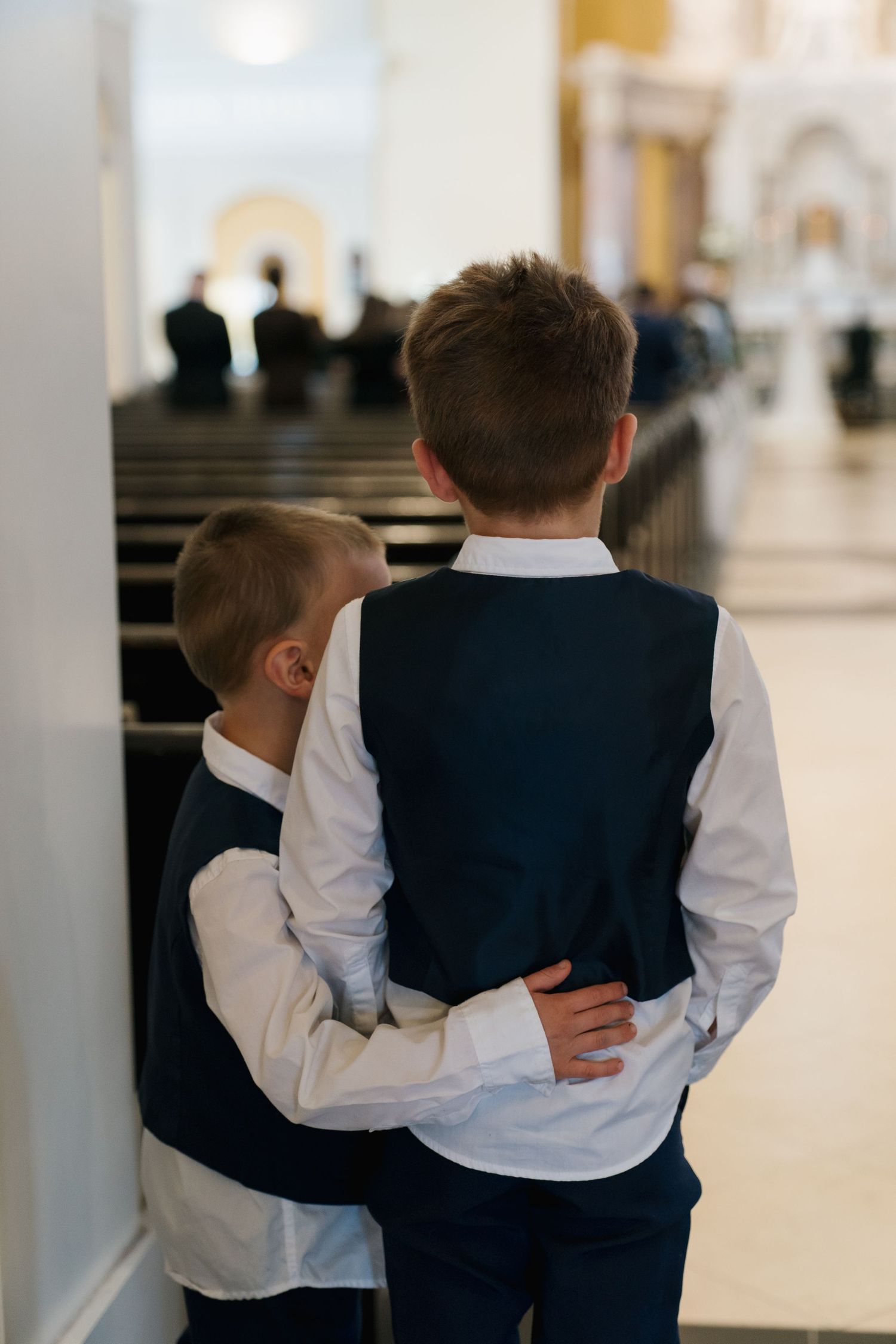 Two young boys in white shirts and navy vests stand together in a church aisle, one embracing the other from behind.