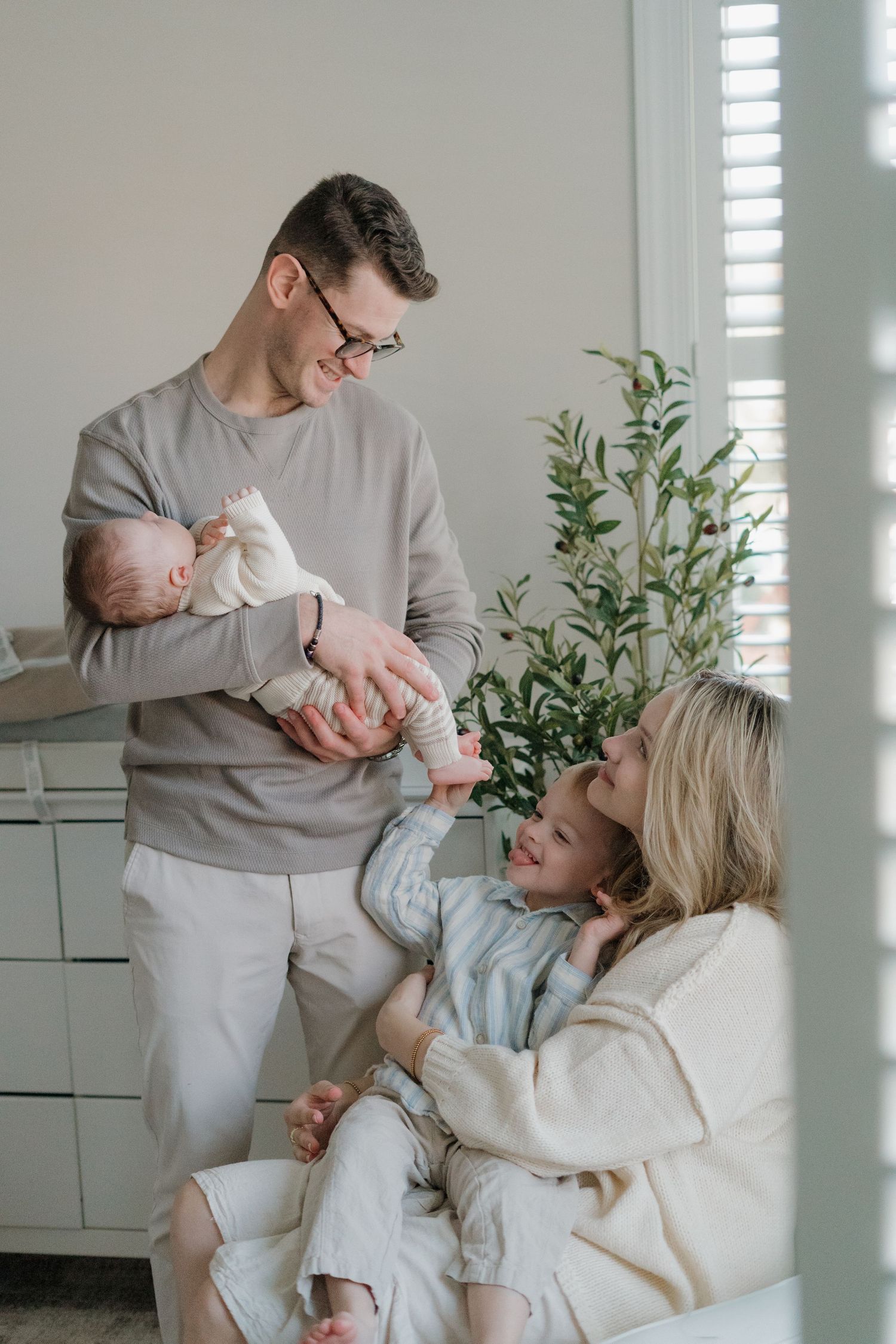 Smiling father holds newborn baby while mother and toddler interact in bright modern home interior.