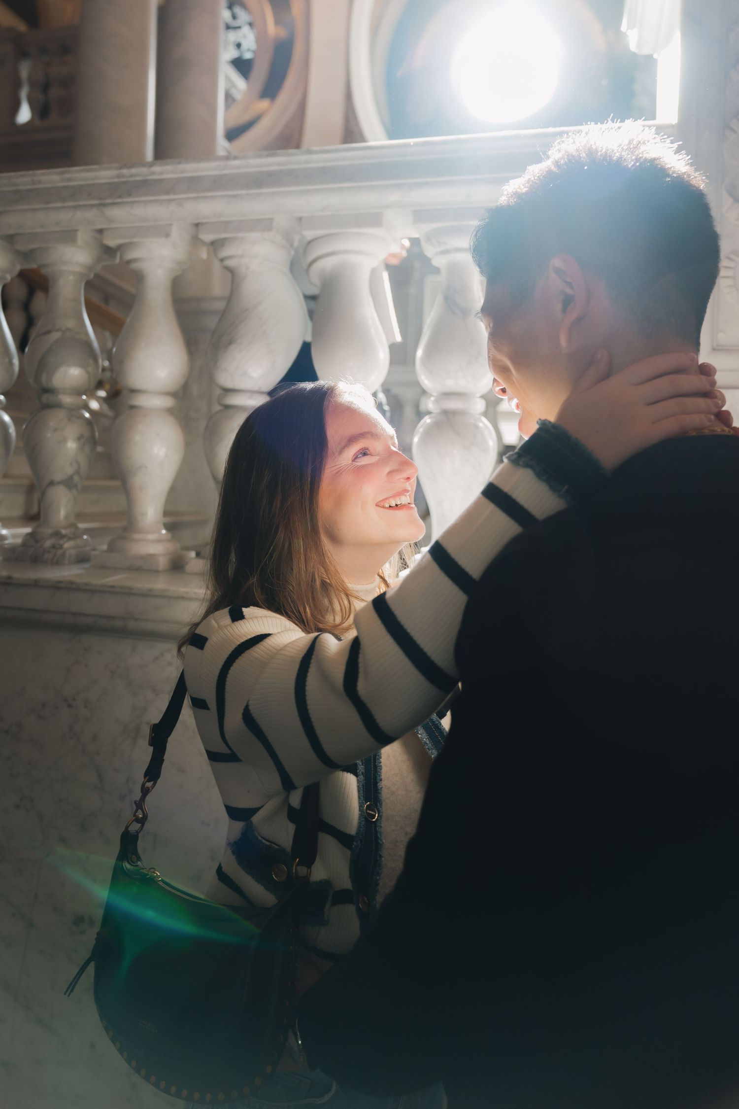 Couple shares a romantic moment near marble balustrade with dramatic backlight in elegant interior.