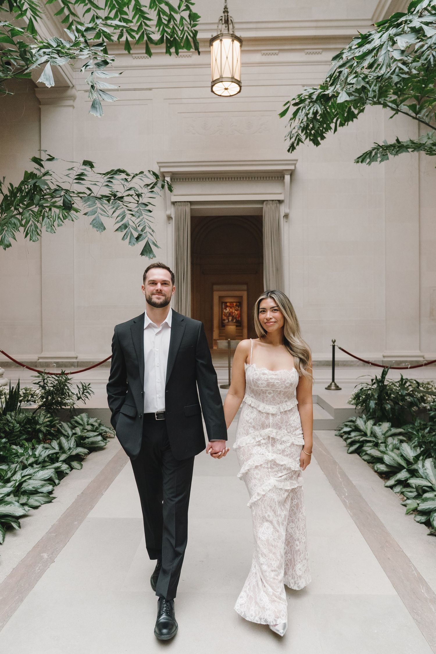 Bride in lace gown and groom walking hand in hand through elegant botanical atrium hallway.