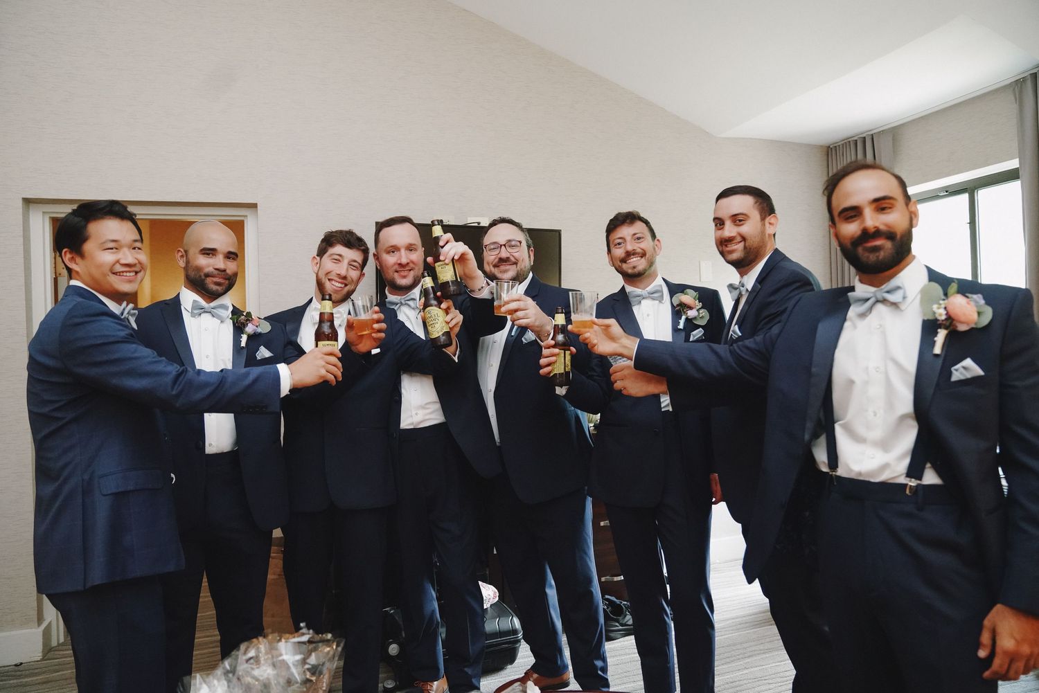A groom and his groomsmen in navy suits toast with drinks indoors before a wedding ceremony.