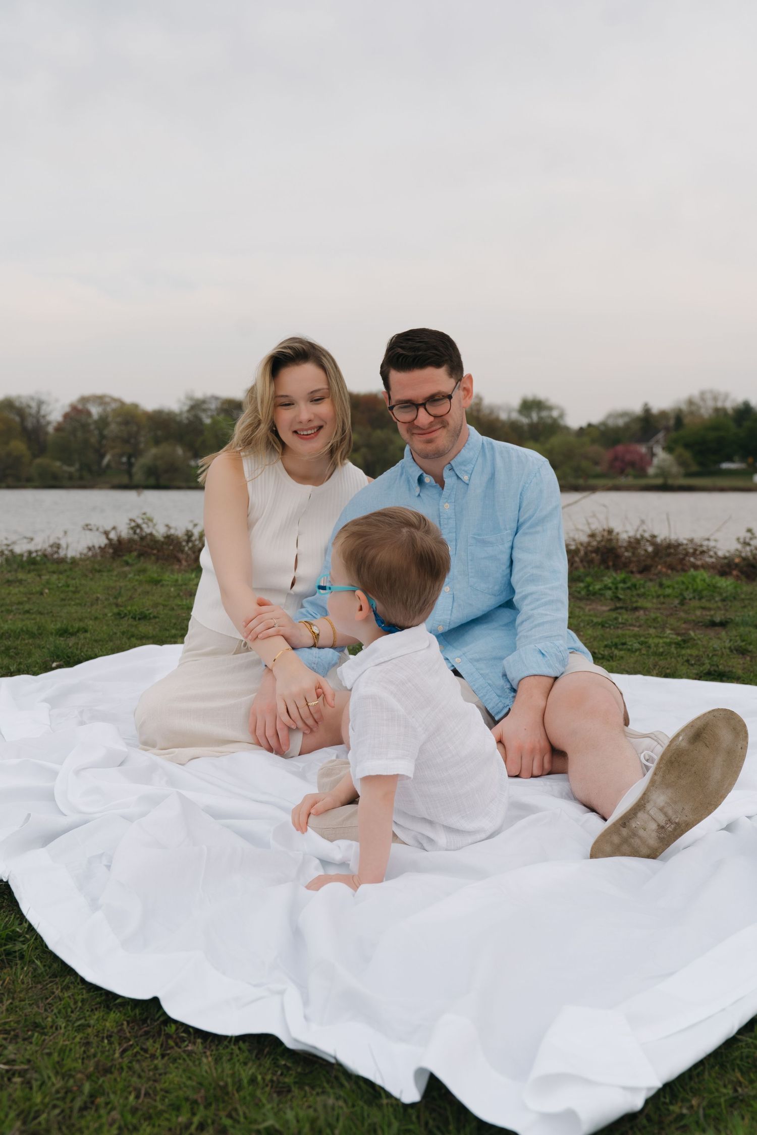 Young couple sitting on a white blanket by a lake, smiling at their toddler child in a white outfit.