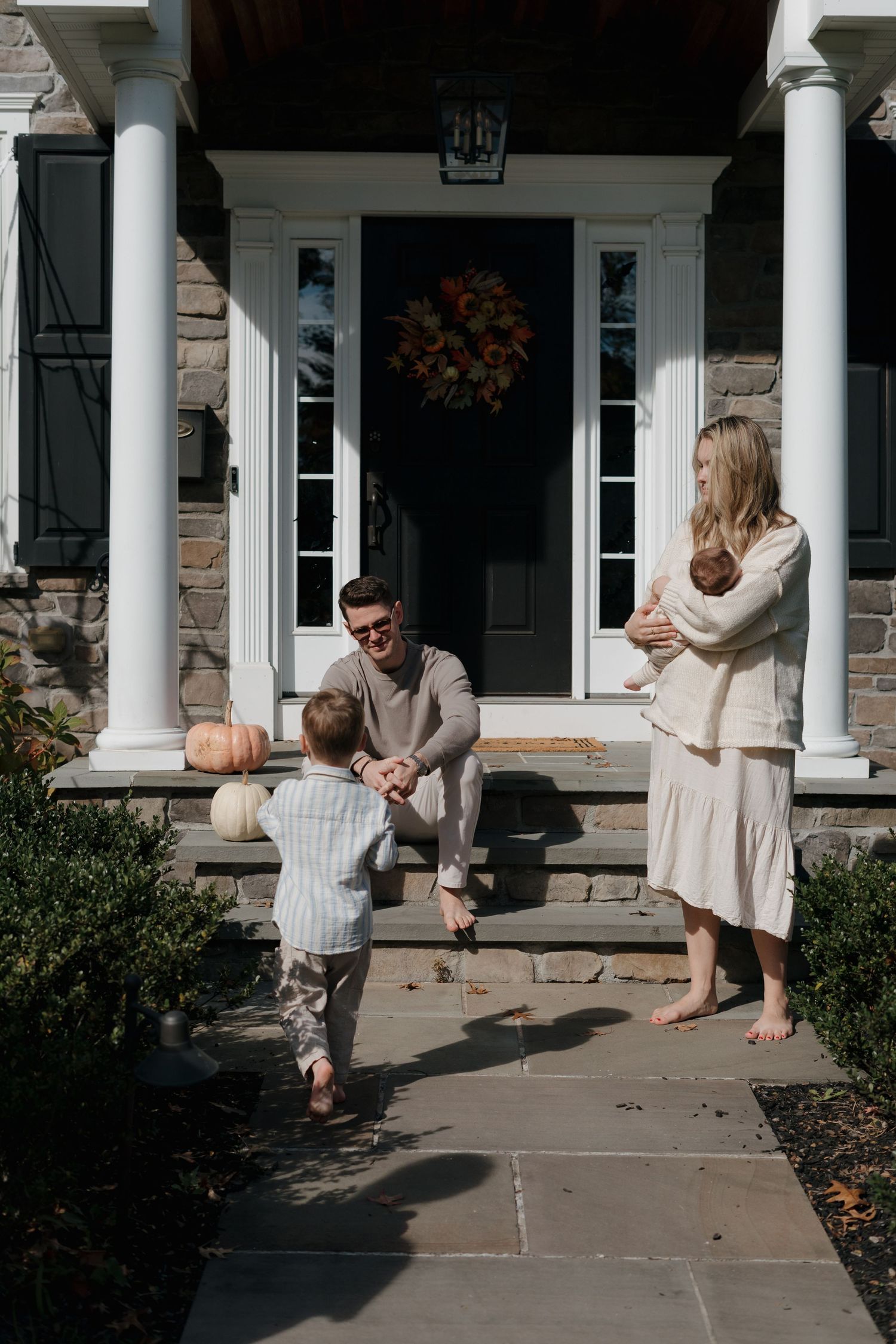 Happy family on home porch steps with father greeting toddler son while mother holds newborn.