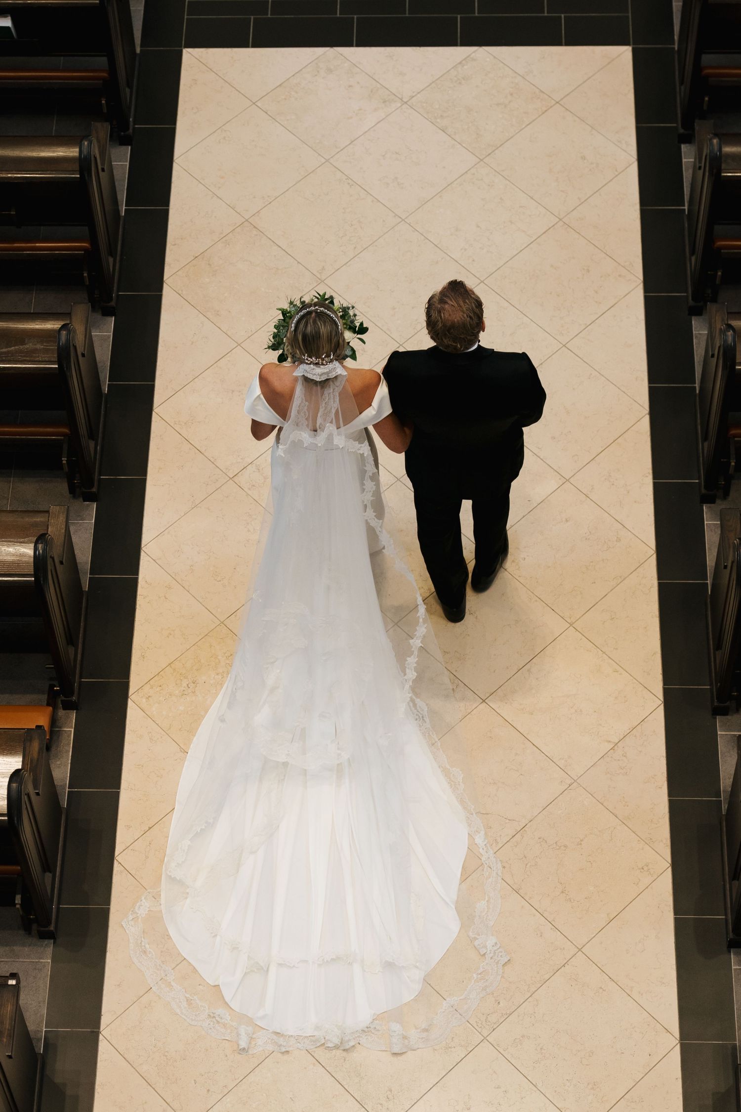 Aerial view of bride in white gown and veil walking down church aisle with escort on wedding day.