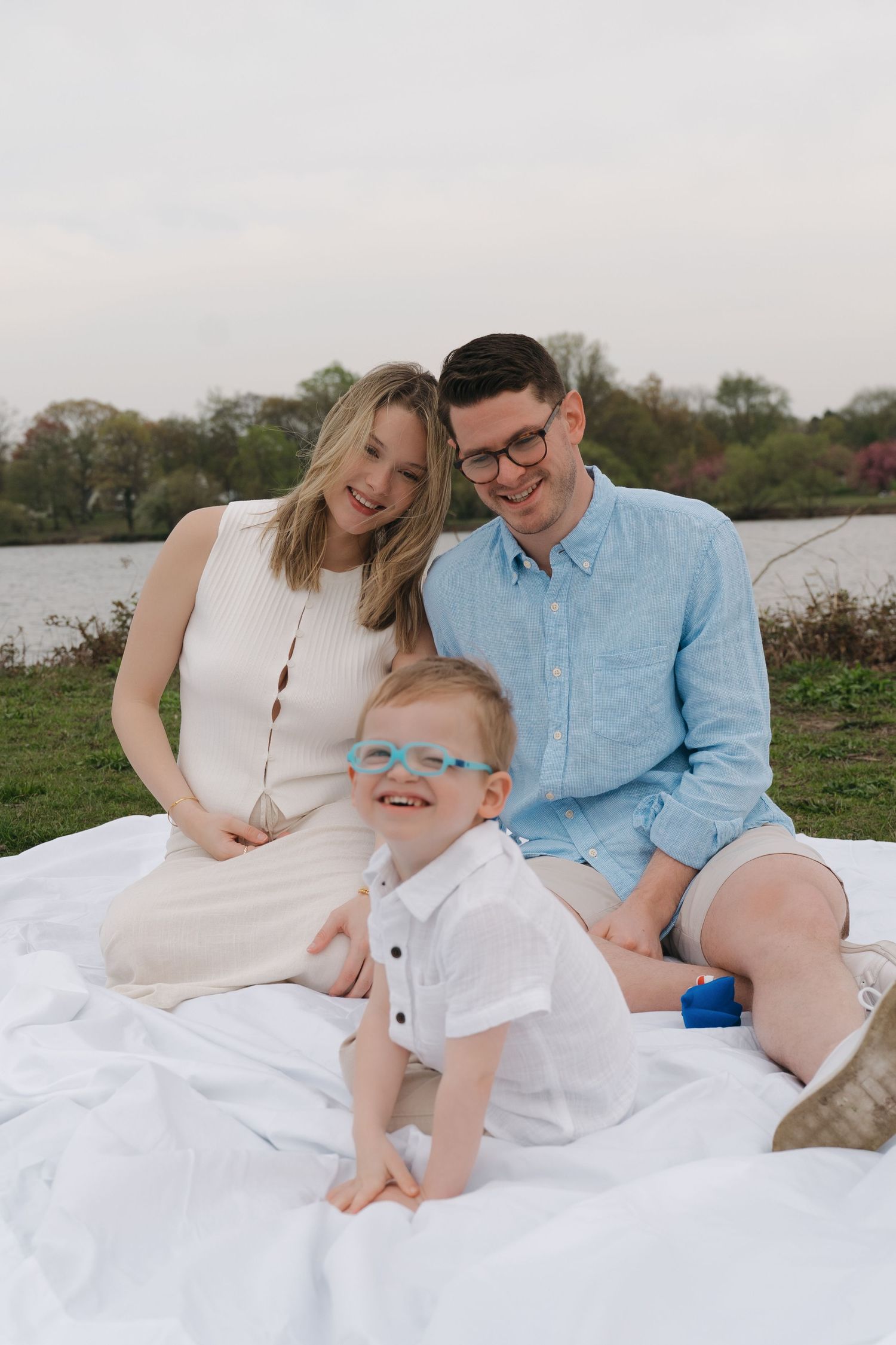 Young family with a toddler boy wearing blue glasses sitting on a white blanket by a lake outdoors.