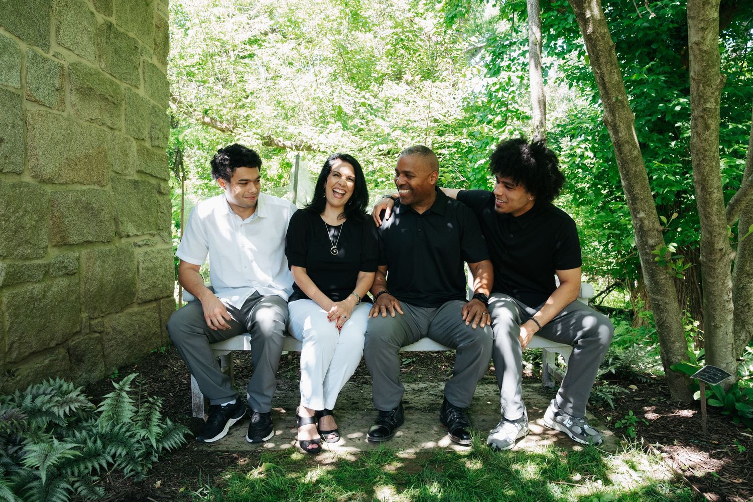 Family of four smiling and sitting together outdoors in lush green garden setting.