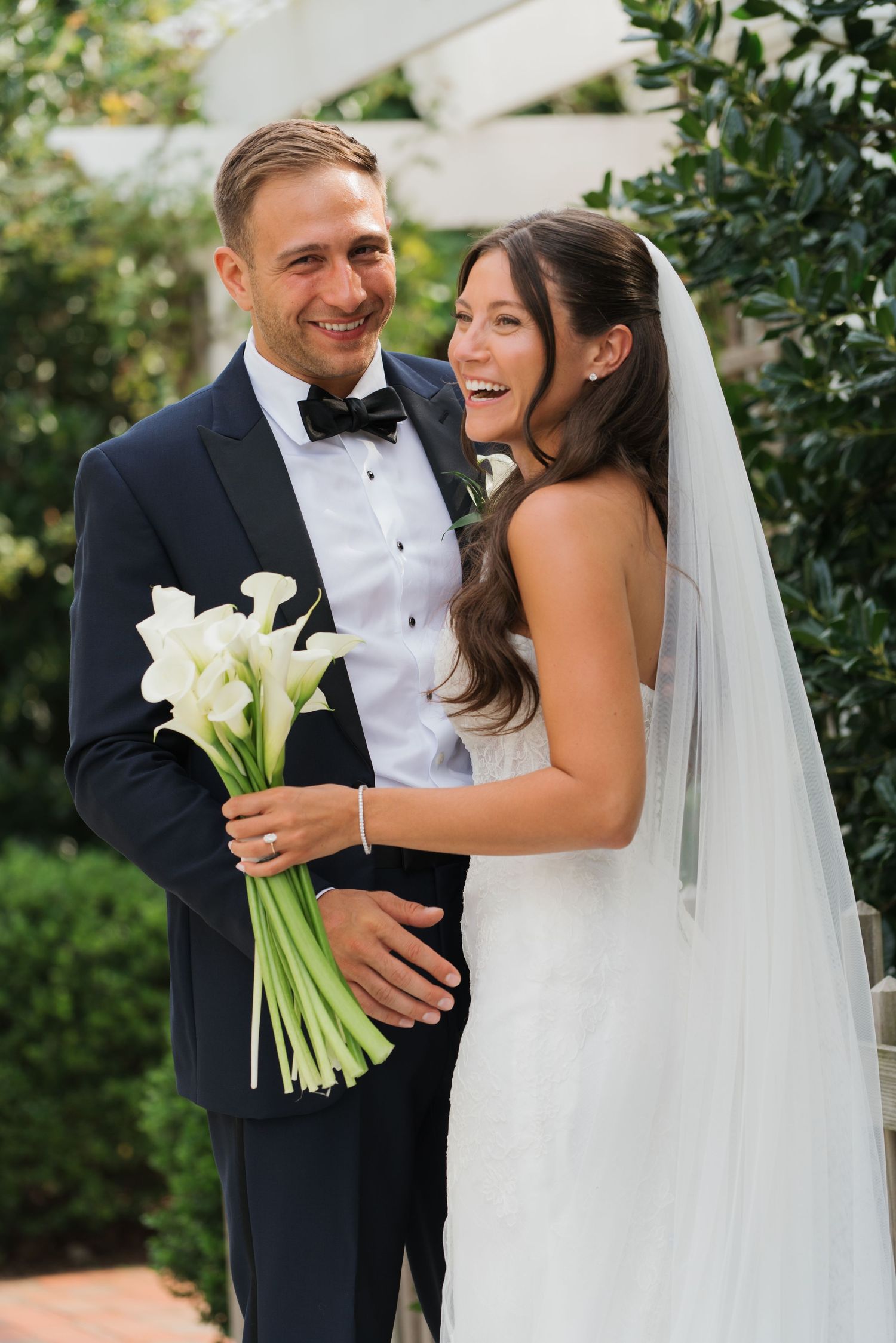 Bride and groom laughing together outdoors, bride holding bouquet, groom in black tuxedo with bow tie at Chesapeake Bay Beach Club in MD