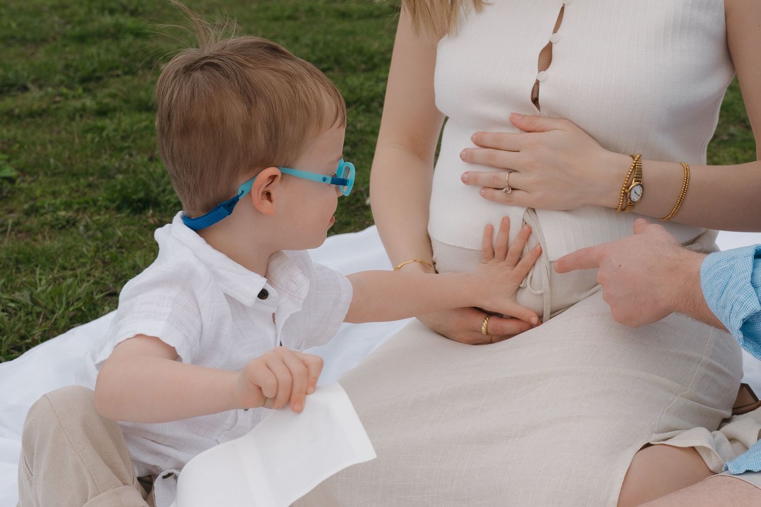 Pregnant woman sitting on grass with young boy in blue glasses touching her baby bump outdoors.
