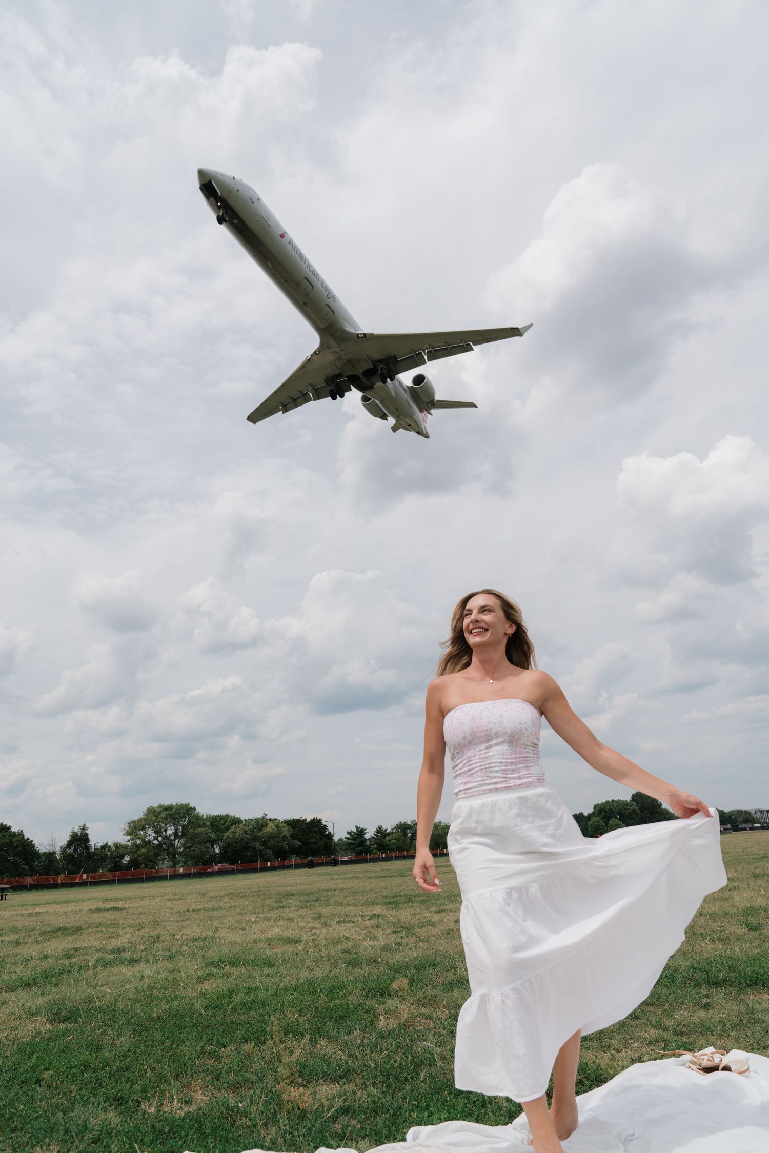 Woman in white dress smiling outdoors as a large airplane flies low overhead against a cloudy sky.