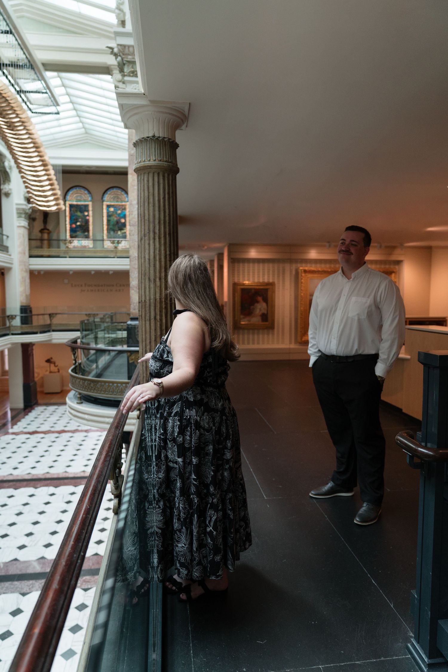 A couple stands on a balcony overlooking an elegant museum atrium with classical columns and skylight.