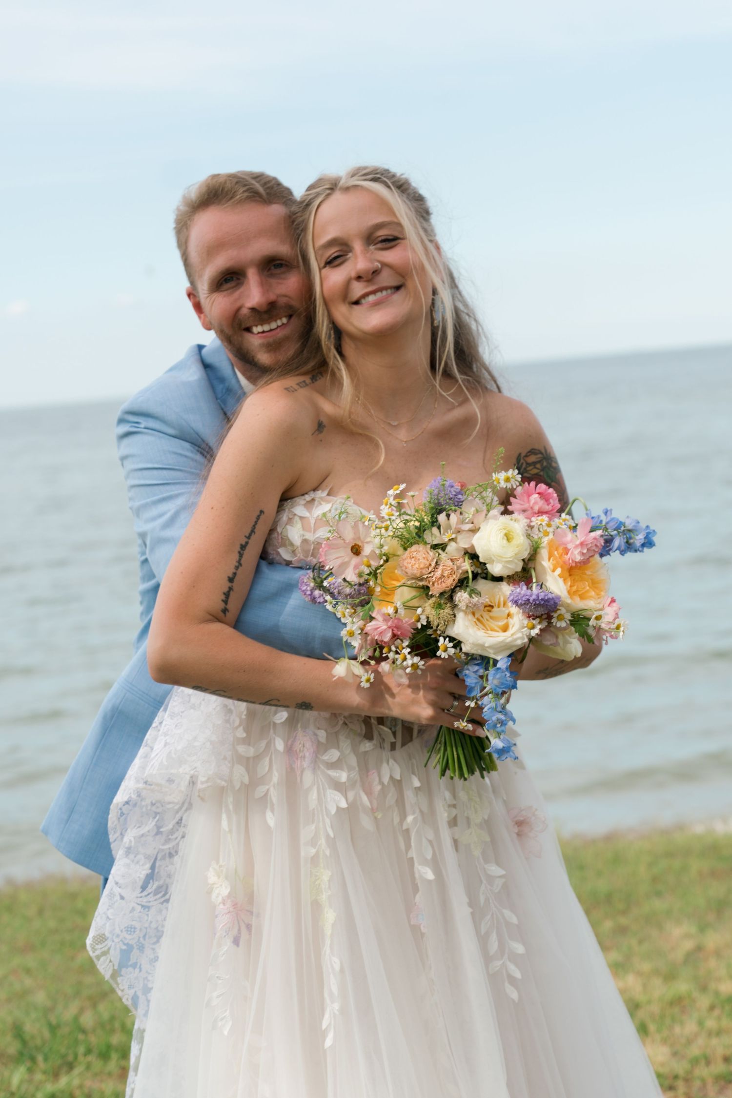 Smiling bride in white floral gown holds colorful bouquet as groom in blue suit embraces her by the sea.
