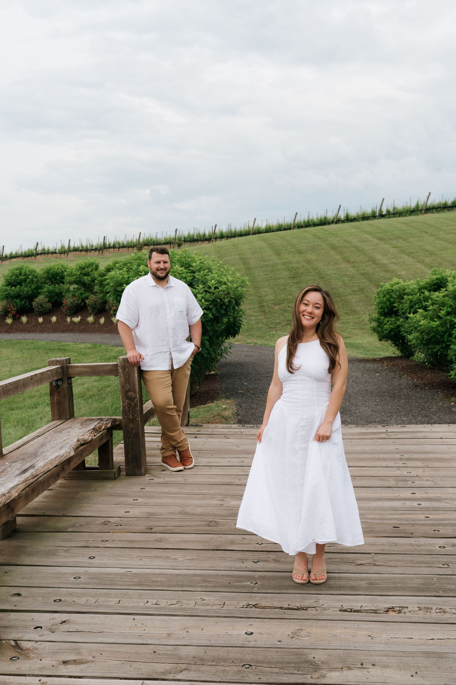Couple in white outfits posing on a wooden bridge at a vineyard, lush green hills in the background.