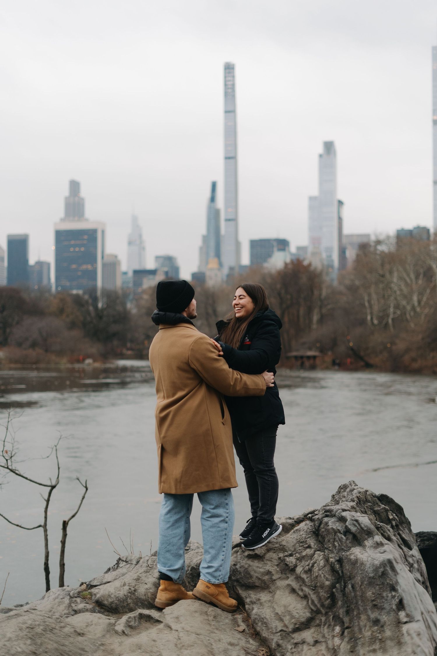 Couple embracing on rocks by a lake with the Manhattan skyline in the background.