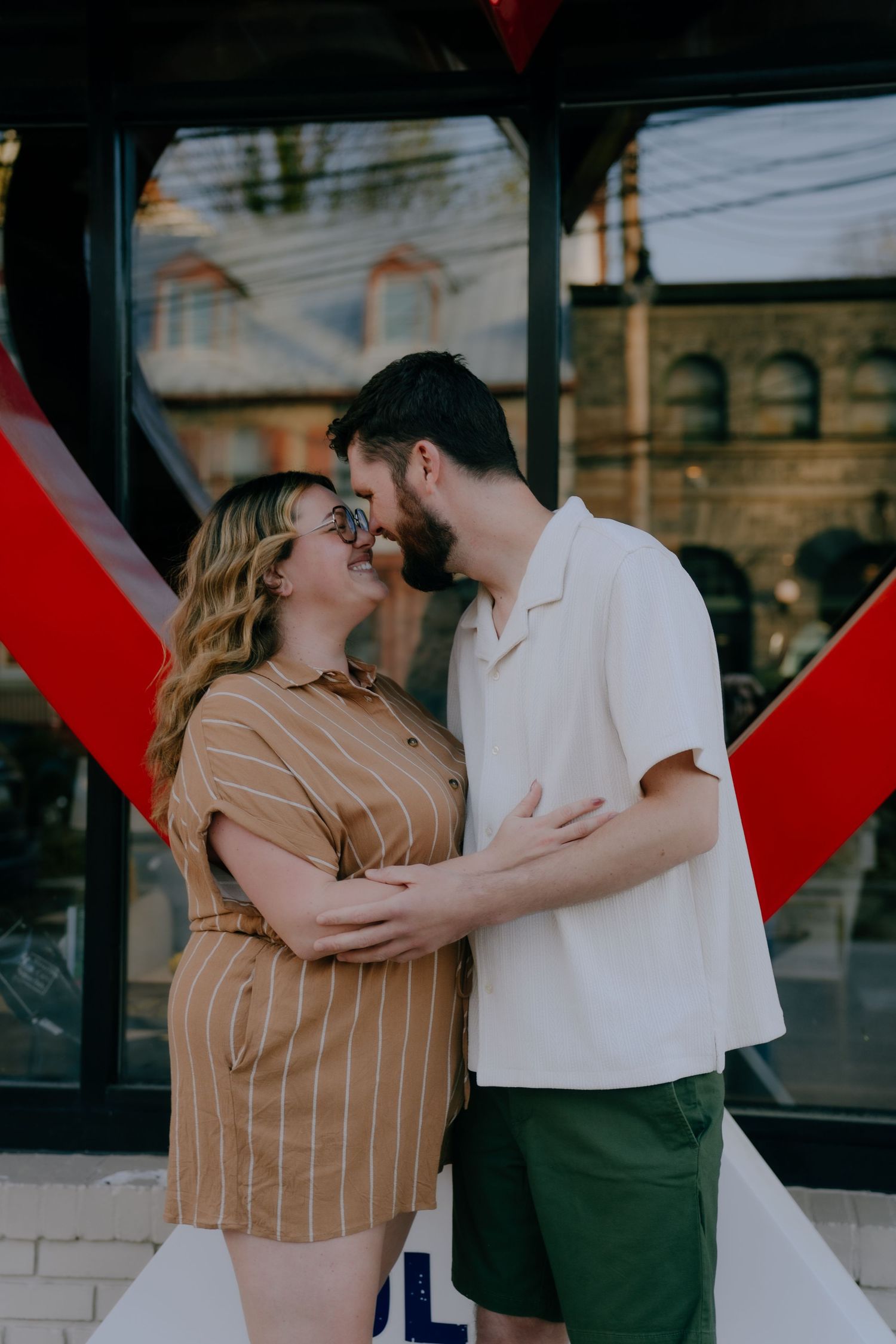 Happy couple sharing a kiss while embracing outside a building with a bold red heart decoration.