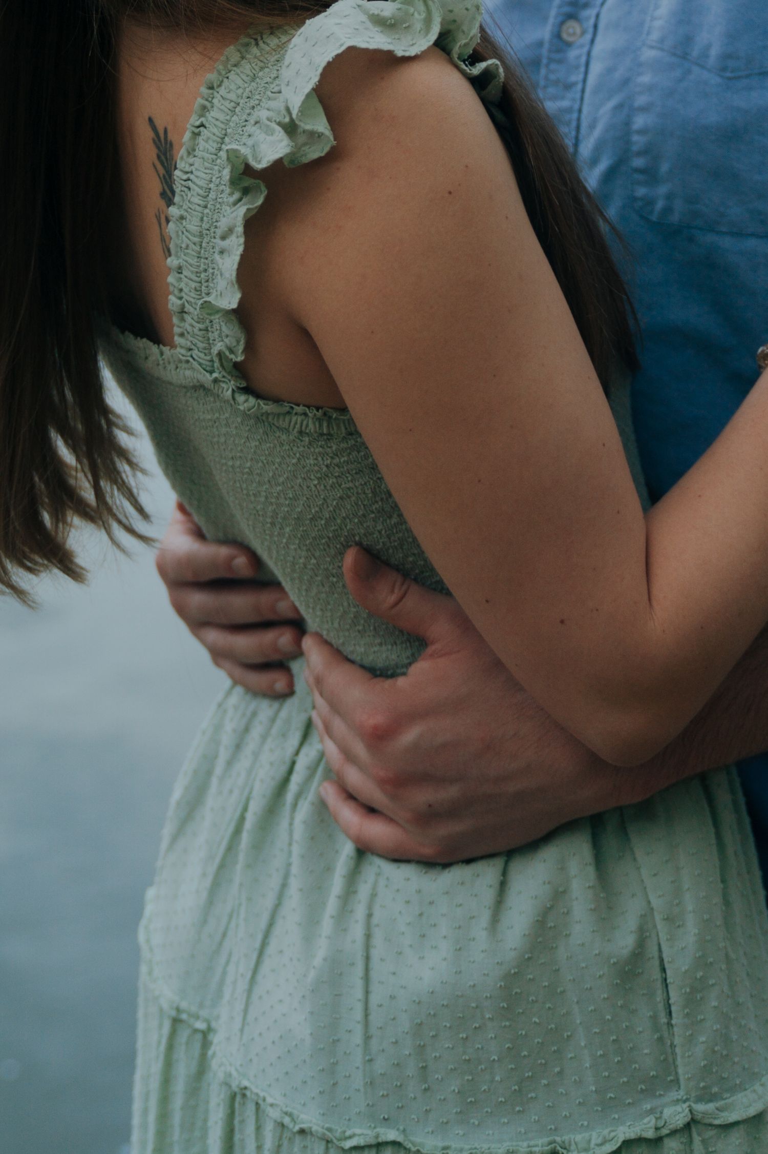 Close-up of couple embracing outdoors, woman in sage green dress with small arrow tattoo visible.