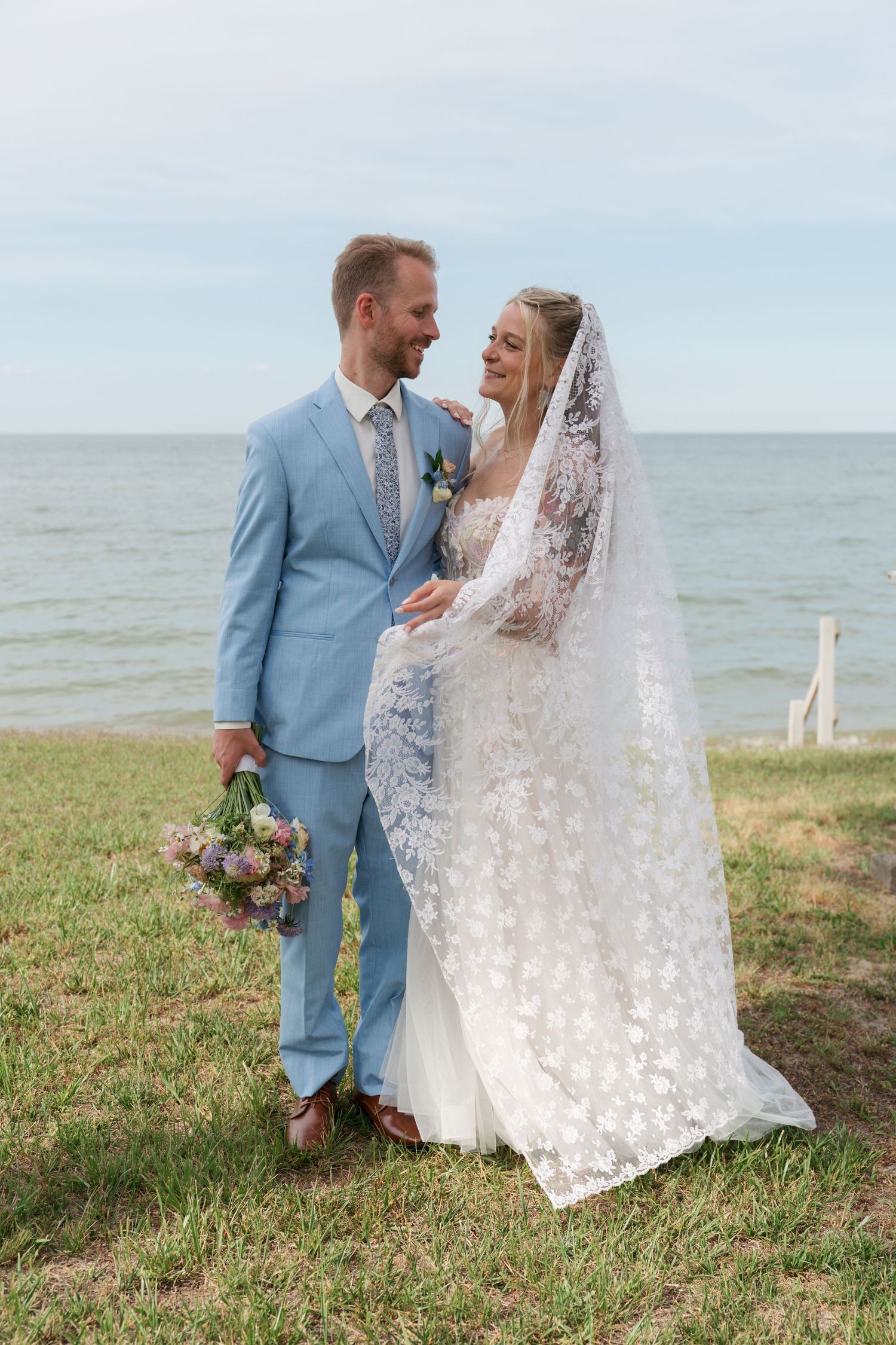 Bride and groom share a romantic moment by the sea, she in lace gown with veil, he in light blue suit holding bouquet.