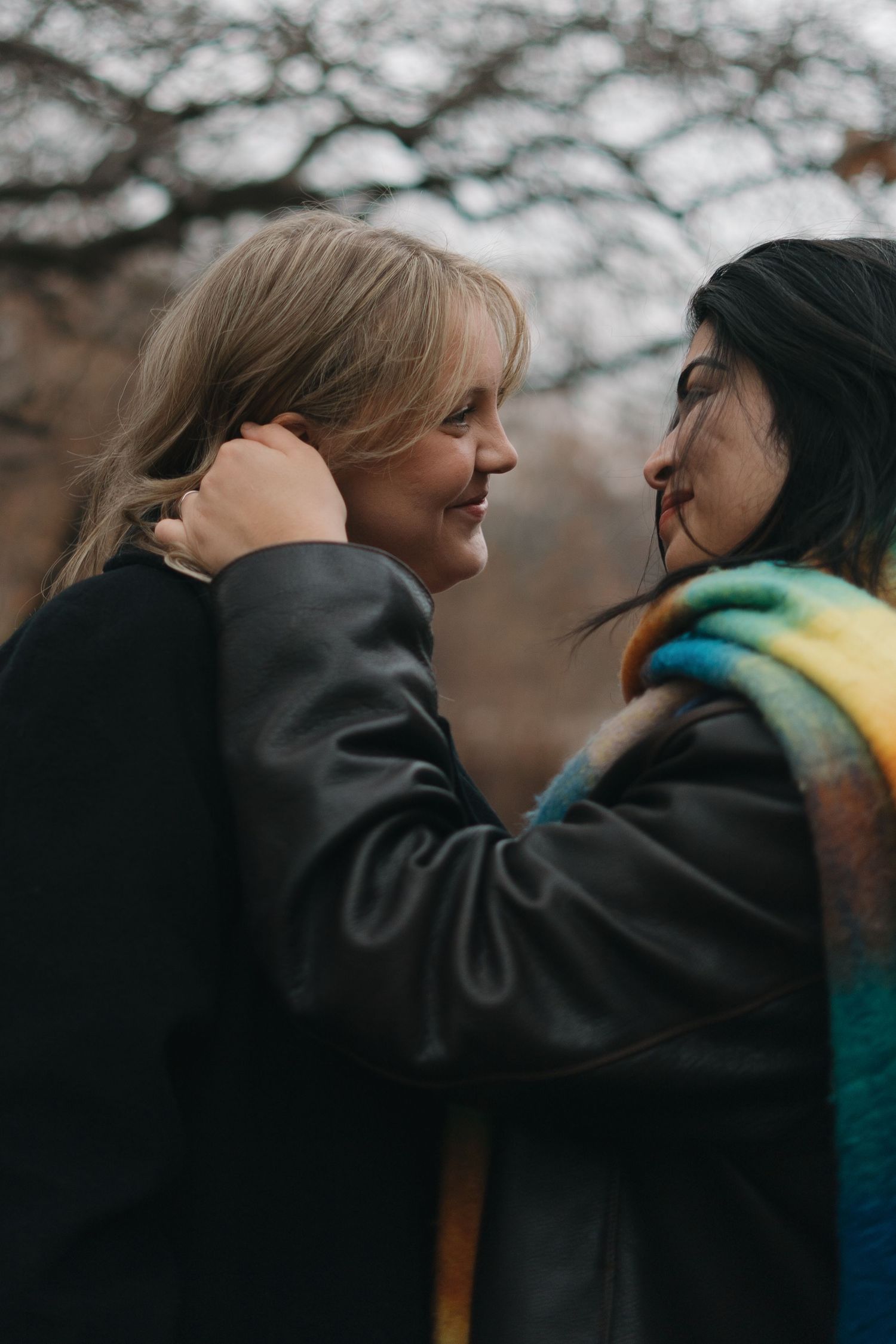 A couple's portrait of two women sharing a moment outdoors, one smiling as the other gently touches her hair in Central Park in NY, NY