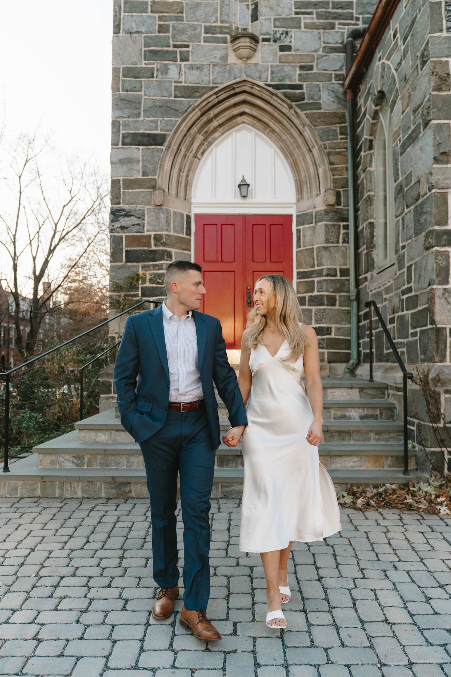 Engaged couple holding hands and walking in front of a stone church with red doors.