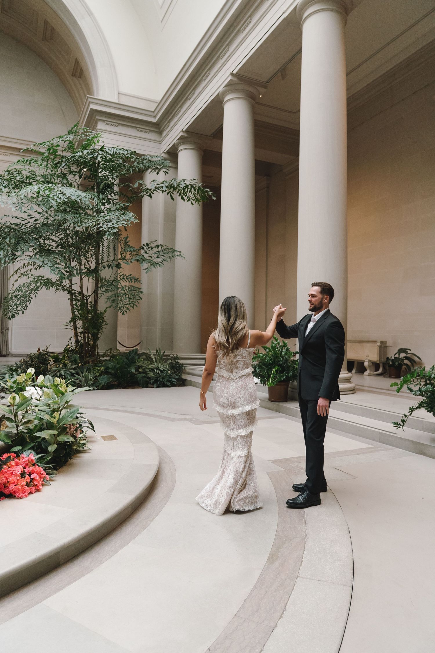 Bride and groom dancing in an elegant museum atrium with tall columns and lush greenery during their wedding reception.