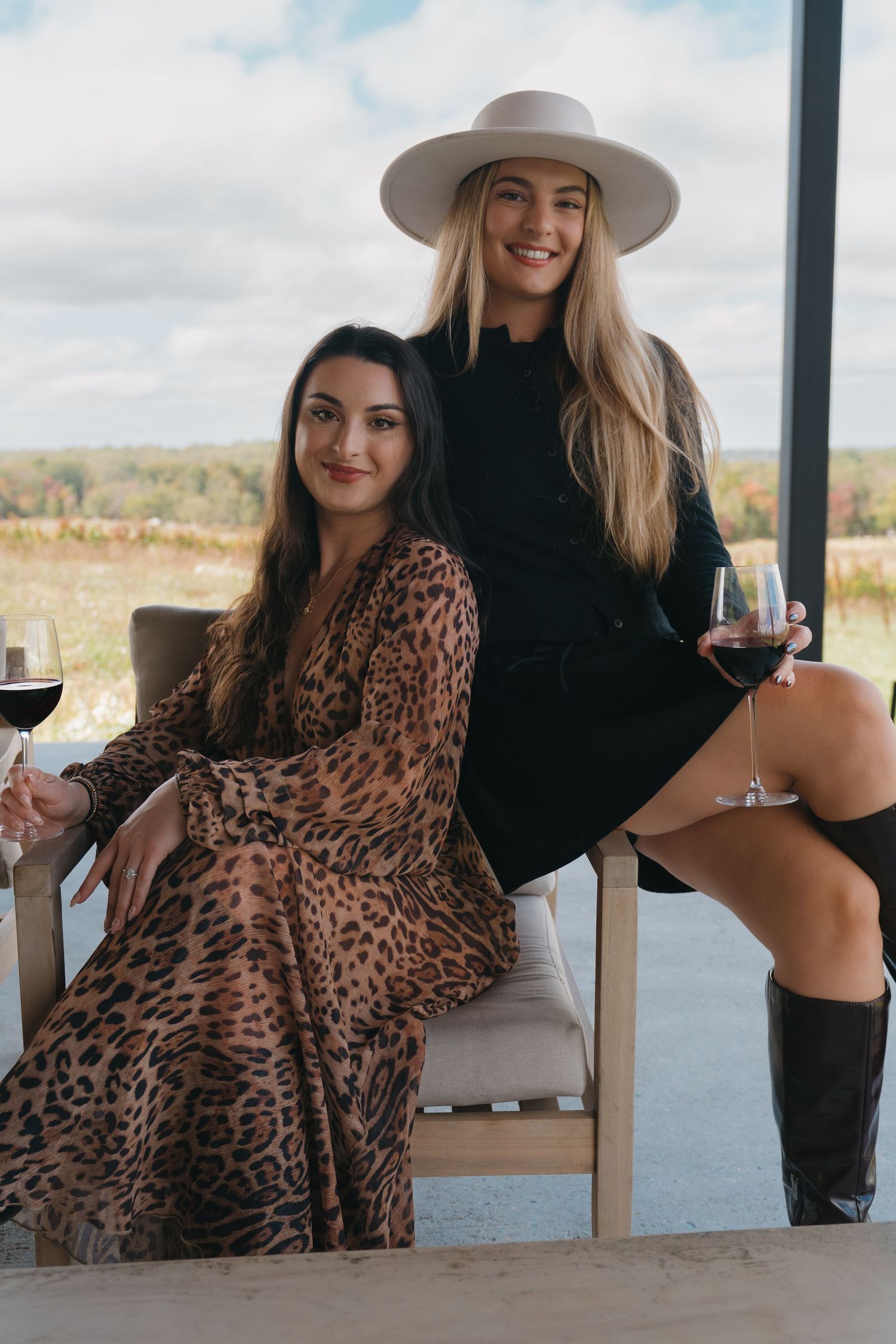 Two women posing with red wine glasses at a vineyard, one in leopard print dress, one in black.