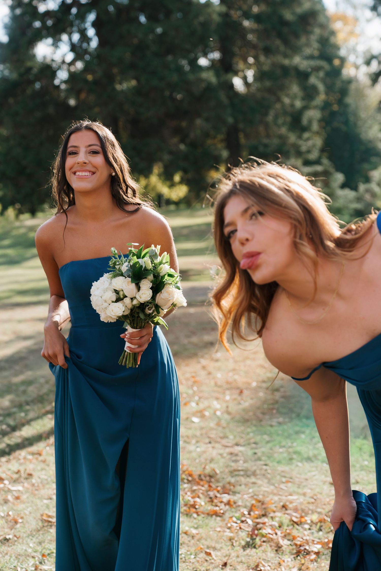 Two bridesmaids in teal gowns pose outdoors, one holding white bouquet smiling, other making playful face.