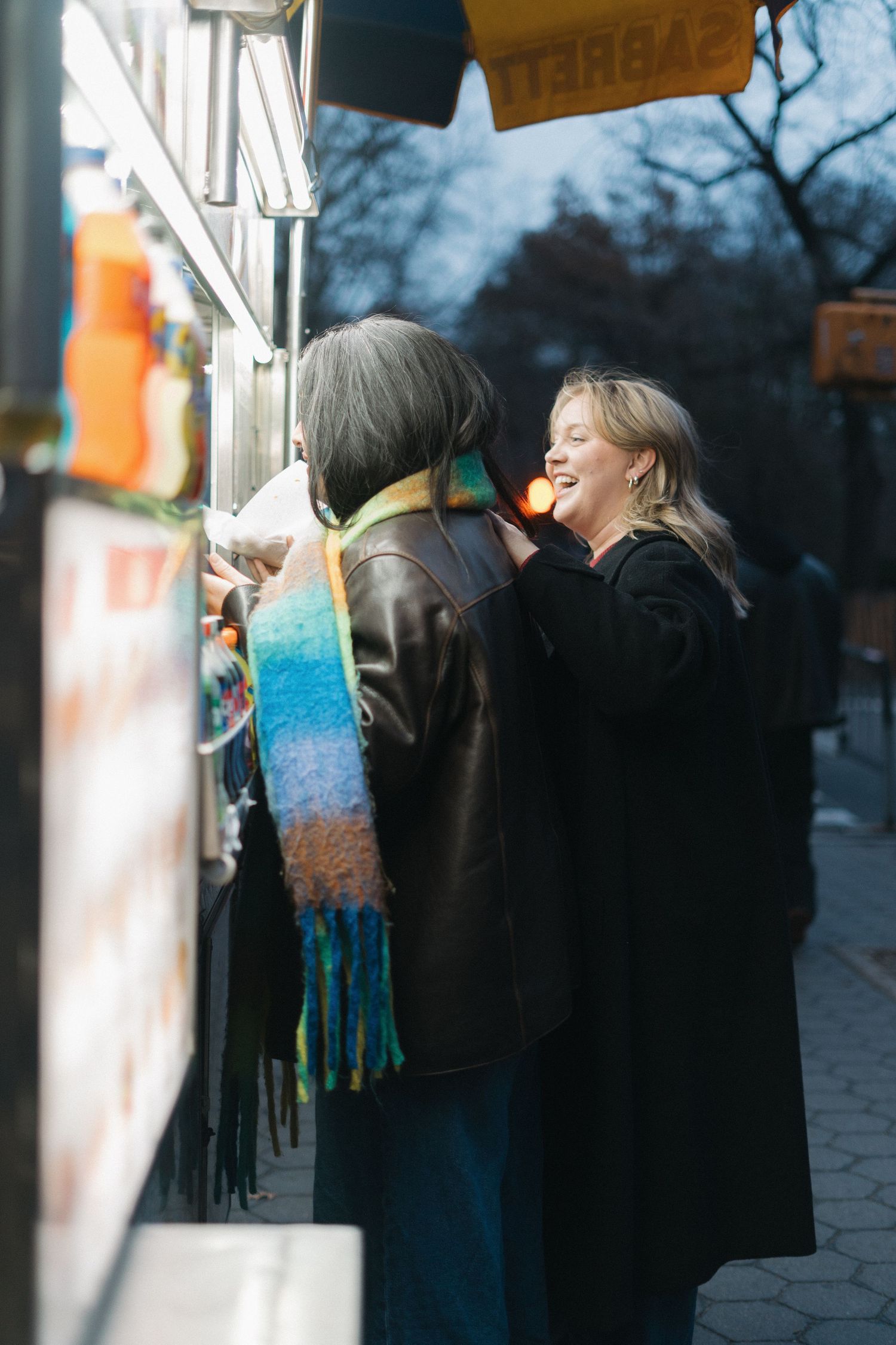 Two women smiling and ordering food at a colorful outdoor street food cart at dusk.