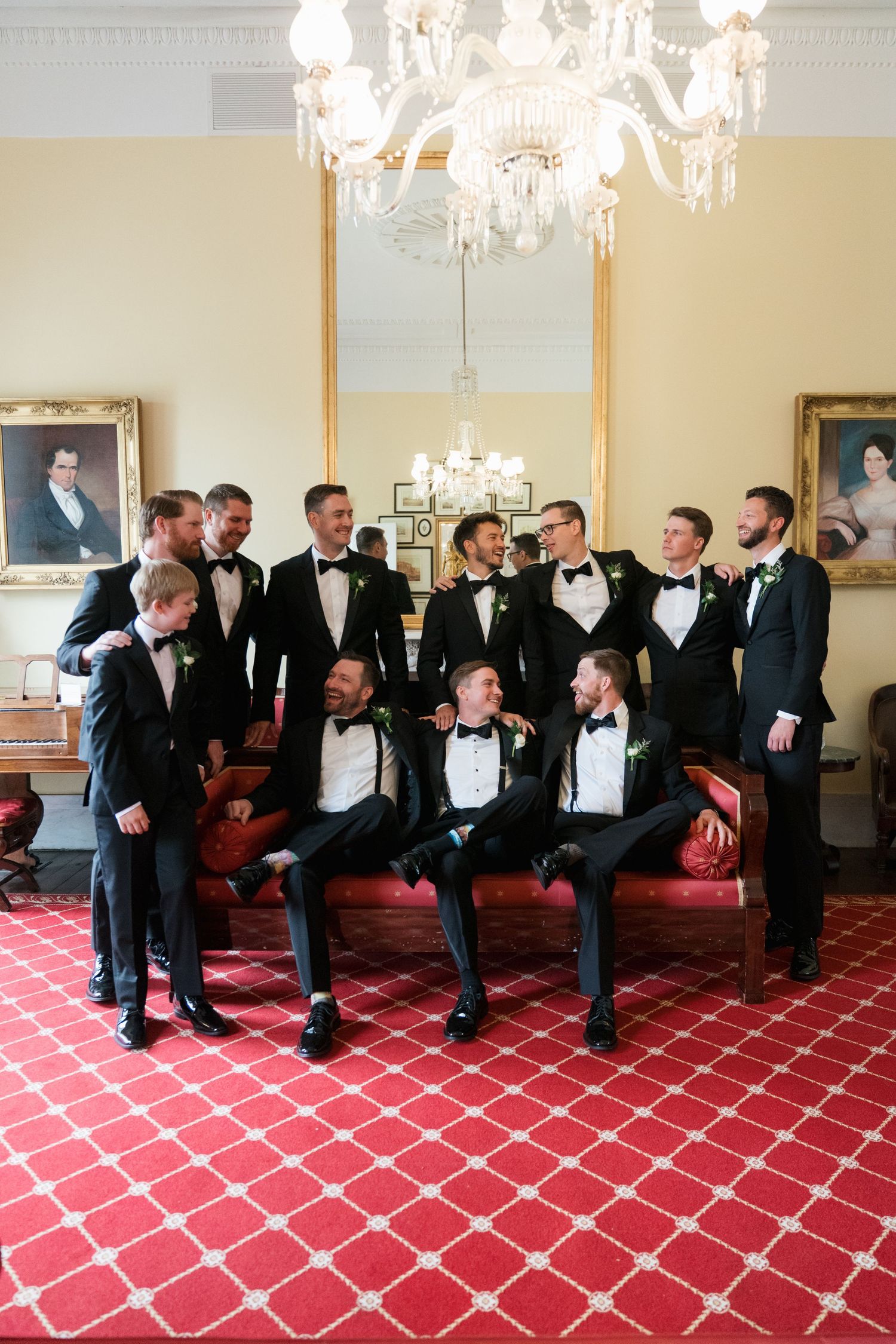 Groomsmen in black tuxedos pose together in an elegant room with red carpet and crystal chandelier.
