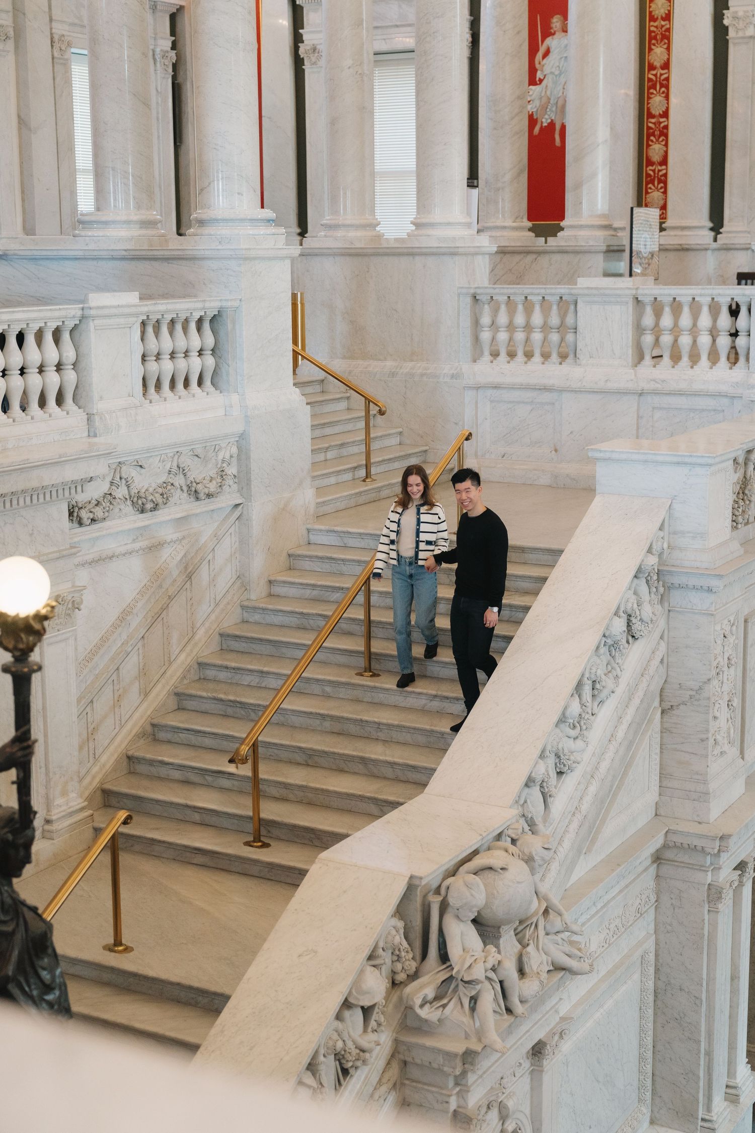Young couple walking down grand marble staircase with gold railings in a historic building.