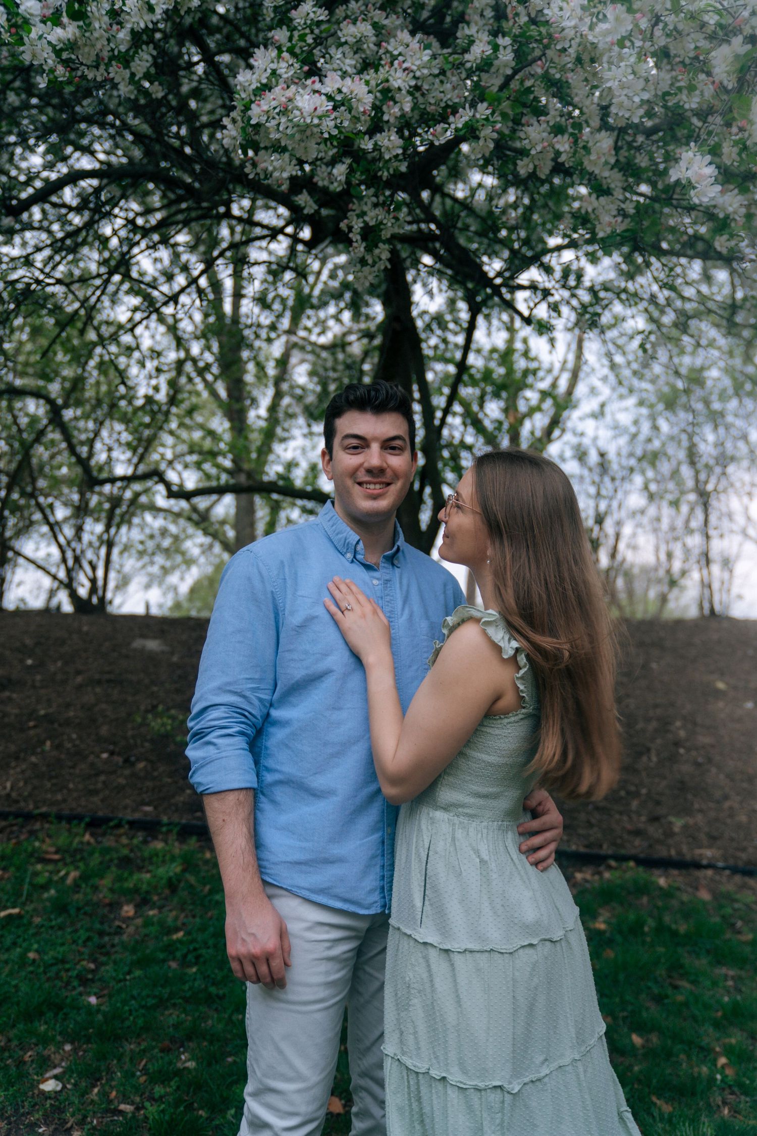 Happy couple standing together under flowering trees, woman in mint dress placing hand on mans chest.