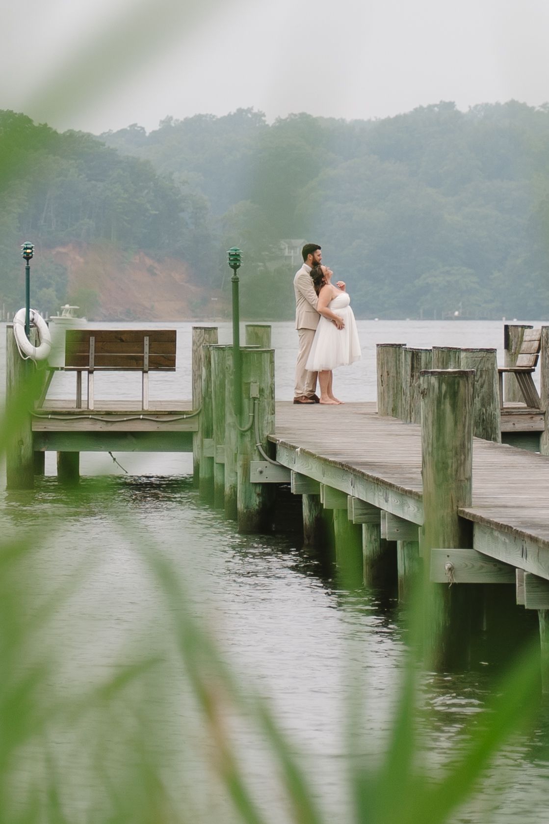 Wedding couple embracing on a misty wooden pier surrounded by green water and lush forested shoreline.