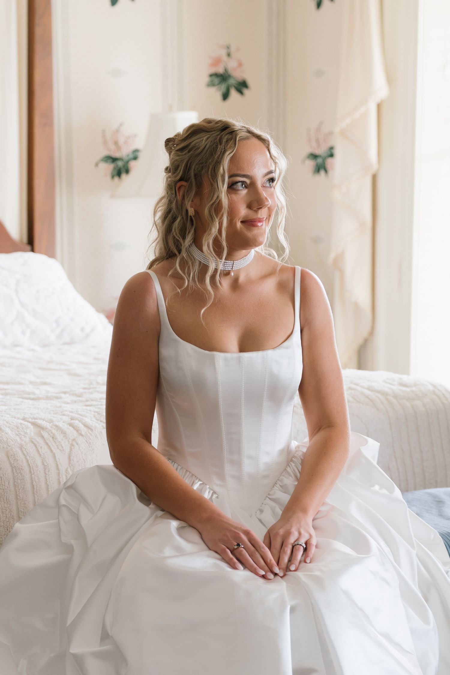 Elegant bride in white satin gown sitting on bed, smiling softly with curly updo hairstyle in bright room.
