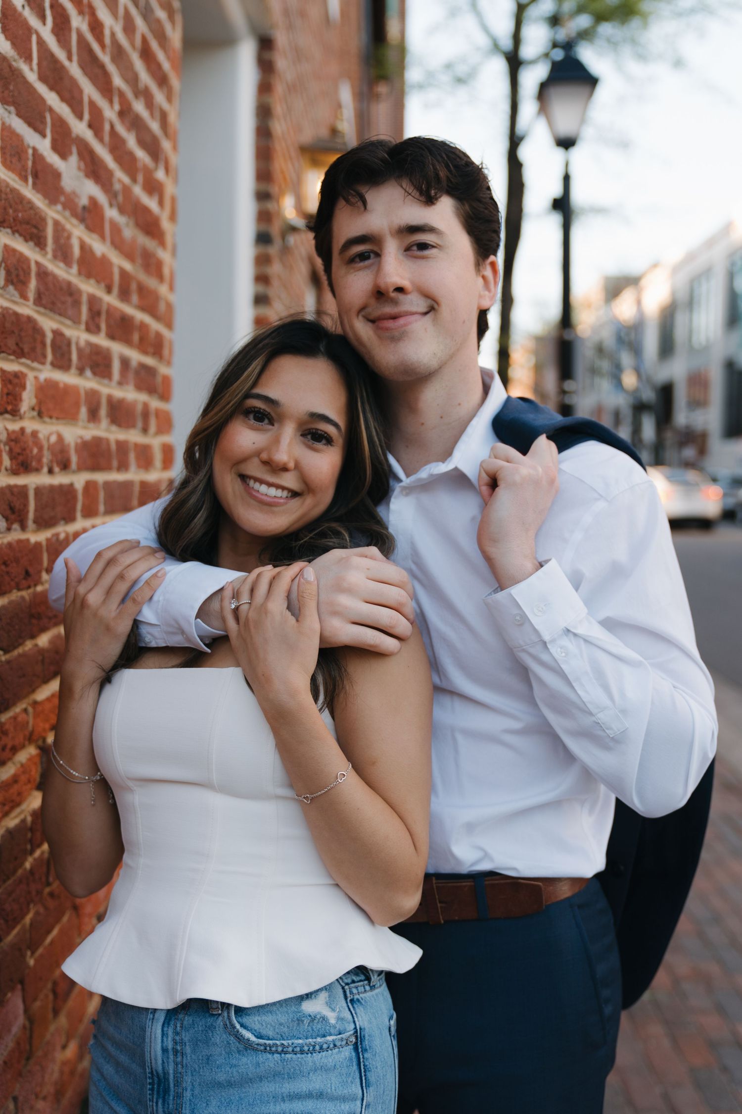 Smiling couple posing together against a brick wall on a charming city street at sunset.