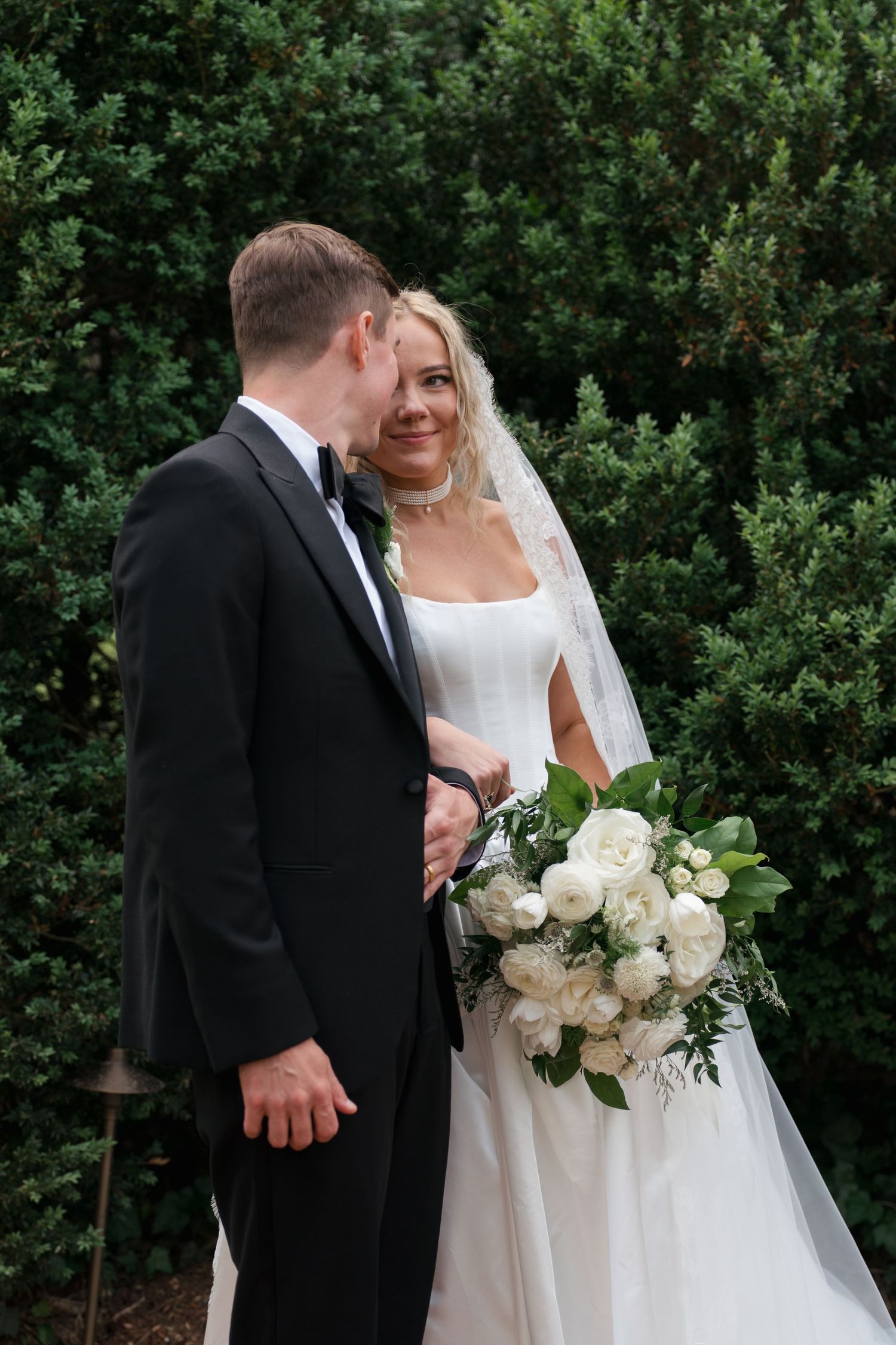 Bride and groom pose together outdoors, she holds white floral bouquet, both dressed in elegant wedding attire before green hedges.