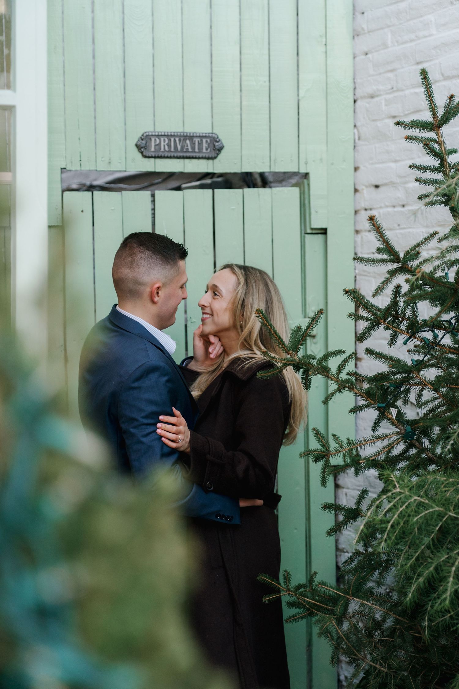 Engaged couple embracing and smiling by a mint green private door with evergreen trees.