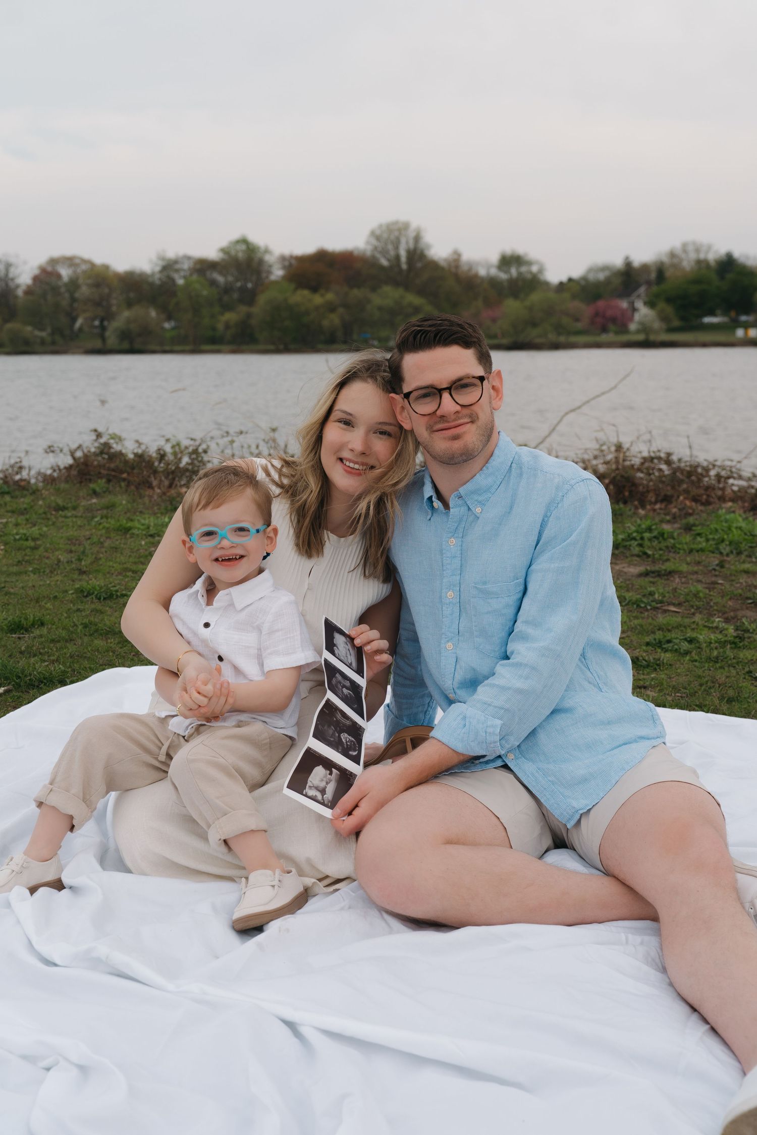 Family sitting on blanket by lake holding ultrasound photos, announcing pregnancy with toddler son.