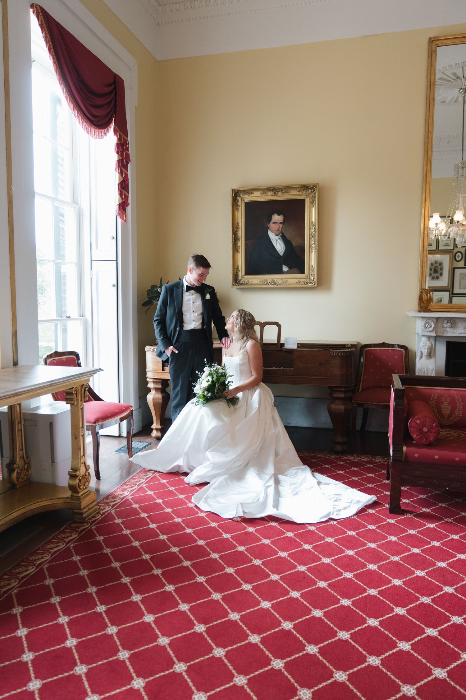 Bride and groom share a romantic moment in an elegant room with red carpet, gold-framed portrait, and crimson curtains.