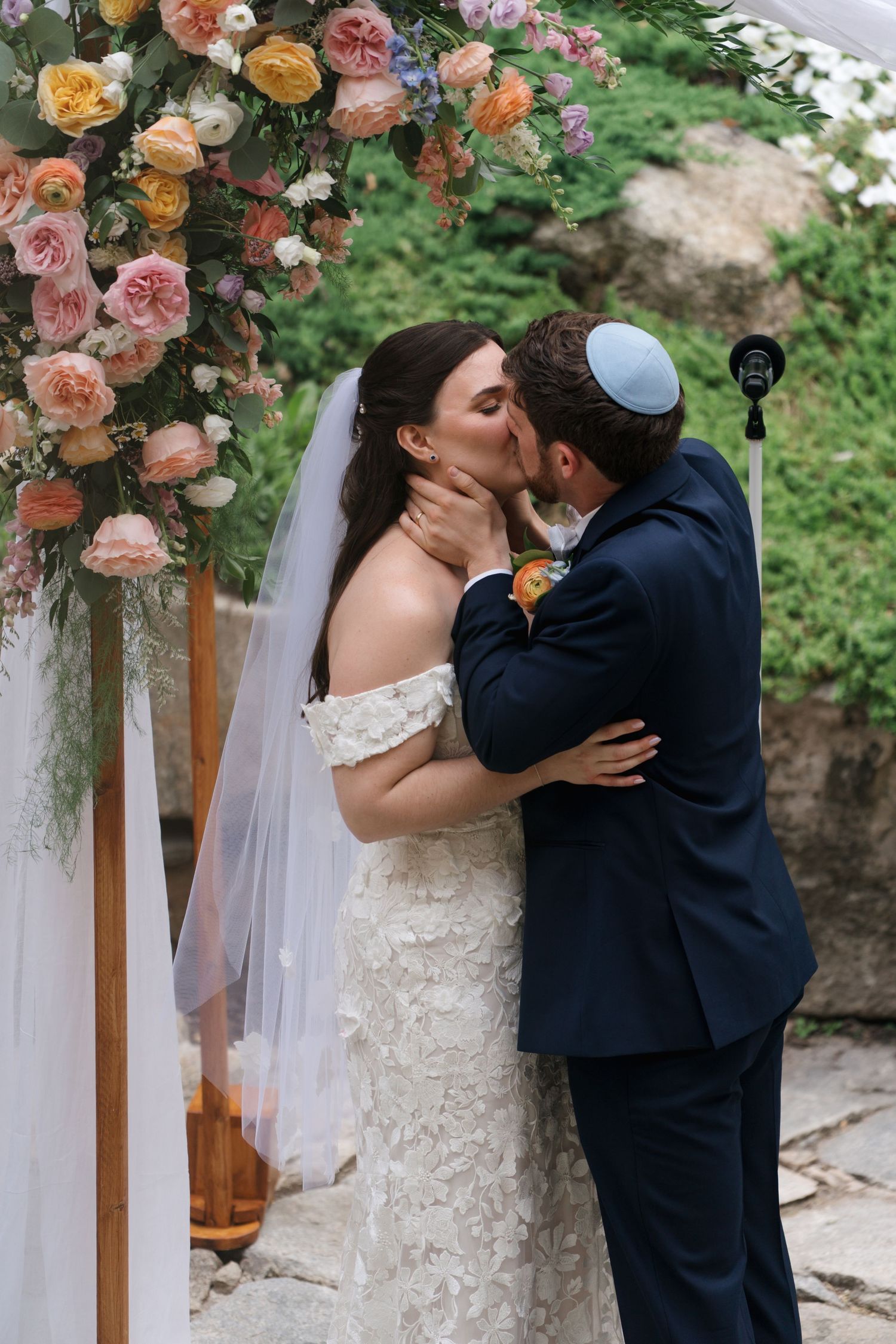 Bride and groom kiss passionately under a vibrant floral chuppah at an outdoor Jewish wedding ceremony.