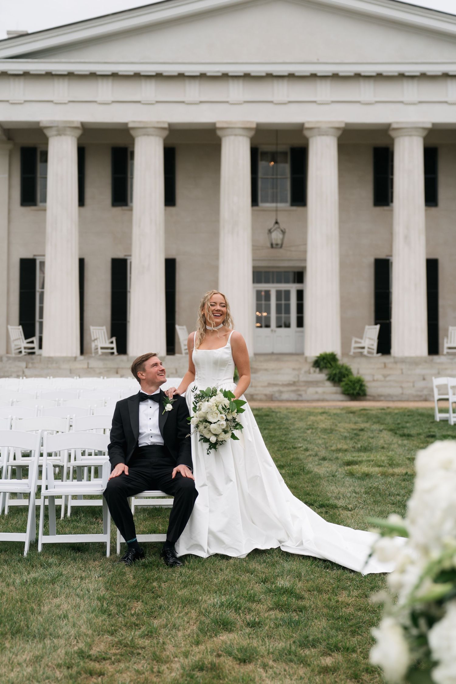 Bride and groom smiling together on lawn of elegant white columned mansion wedding venue with white chairs.