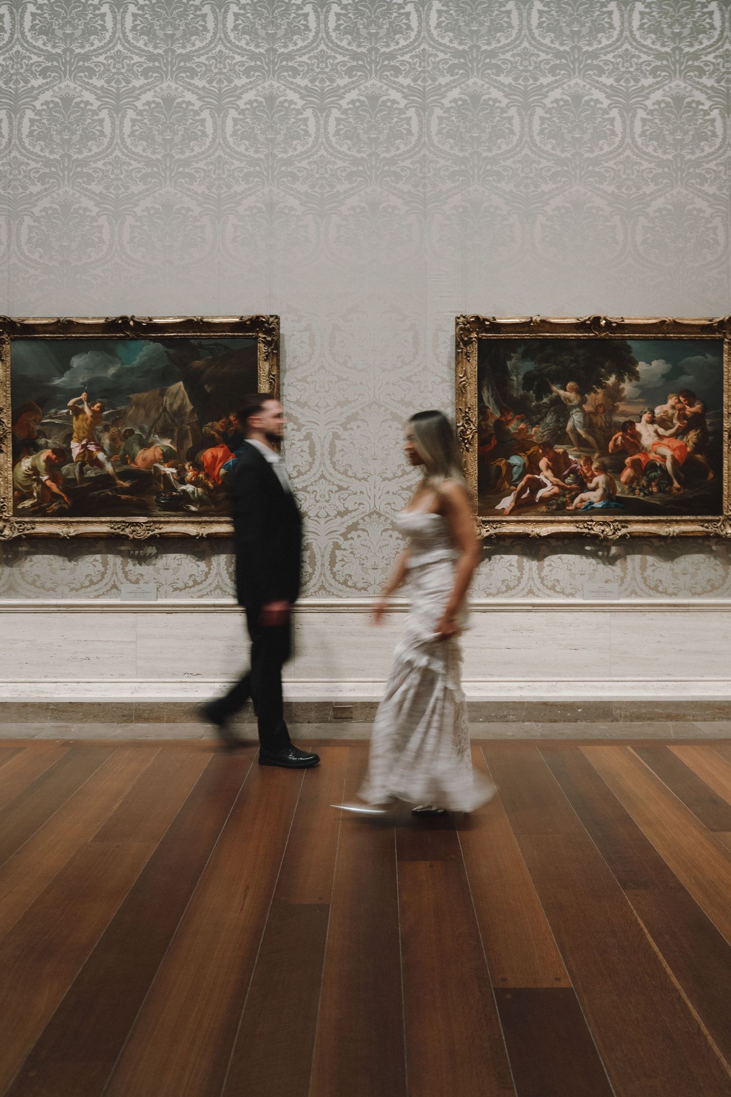 A couple in formal attire walks through an elegant art gallery with classical paintings and ornate wallpaper.