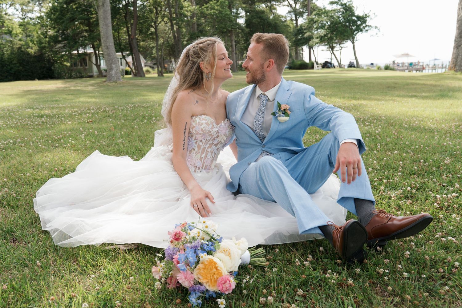 Bride and groom sitting on grass laughing, colorful bouquet nearby, outdoor wedding portrait.