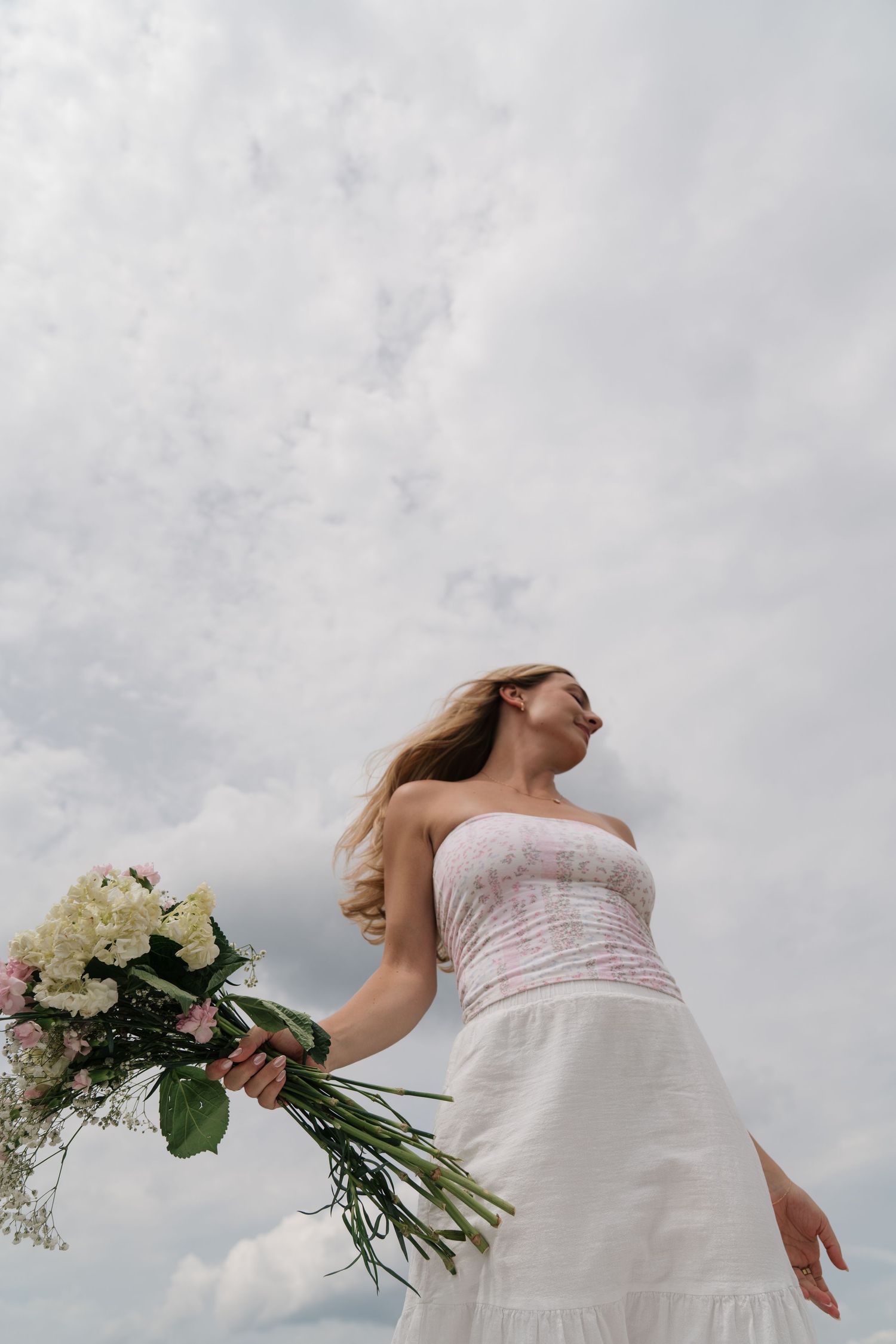 Woman in white strapless dress holding bouquet of flowers against cloudy sky.