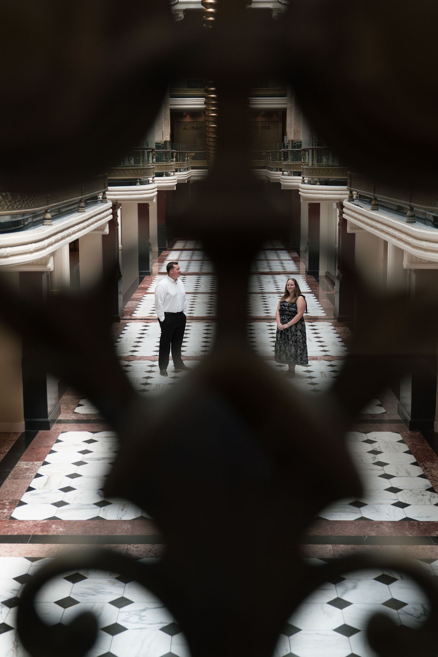 Couple stands in elegant marble-floored atrium viewed through ornate wrought iron railing above.