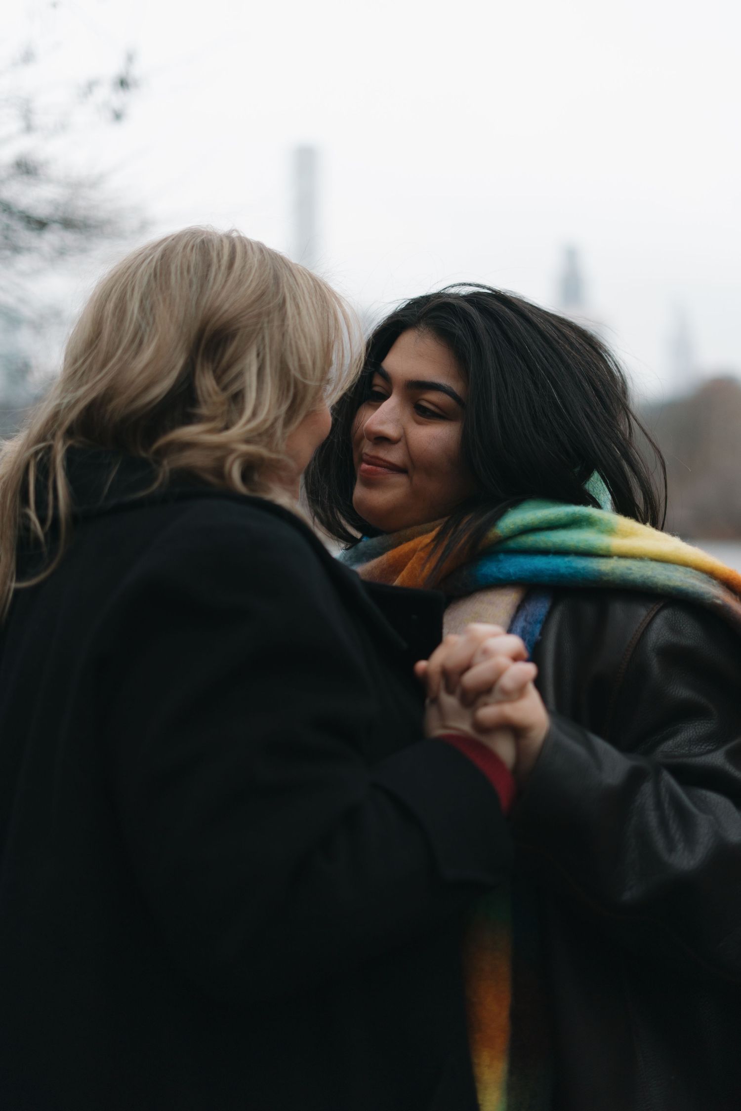 Two women holding hands and sharing a tender moment outdoors with a city skyline behind.