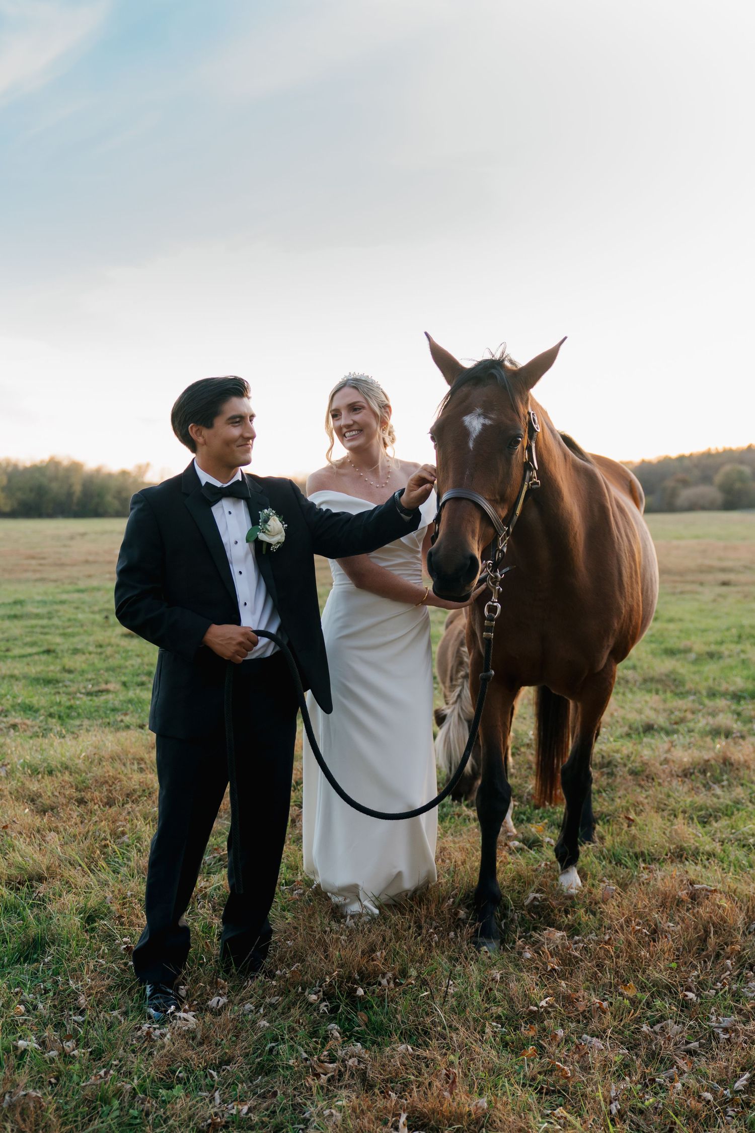 Smiling bride and groom in formal attire standing with a chestnut horse in an open field.