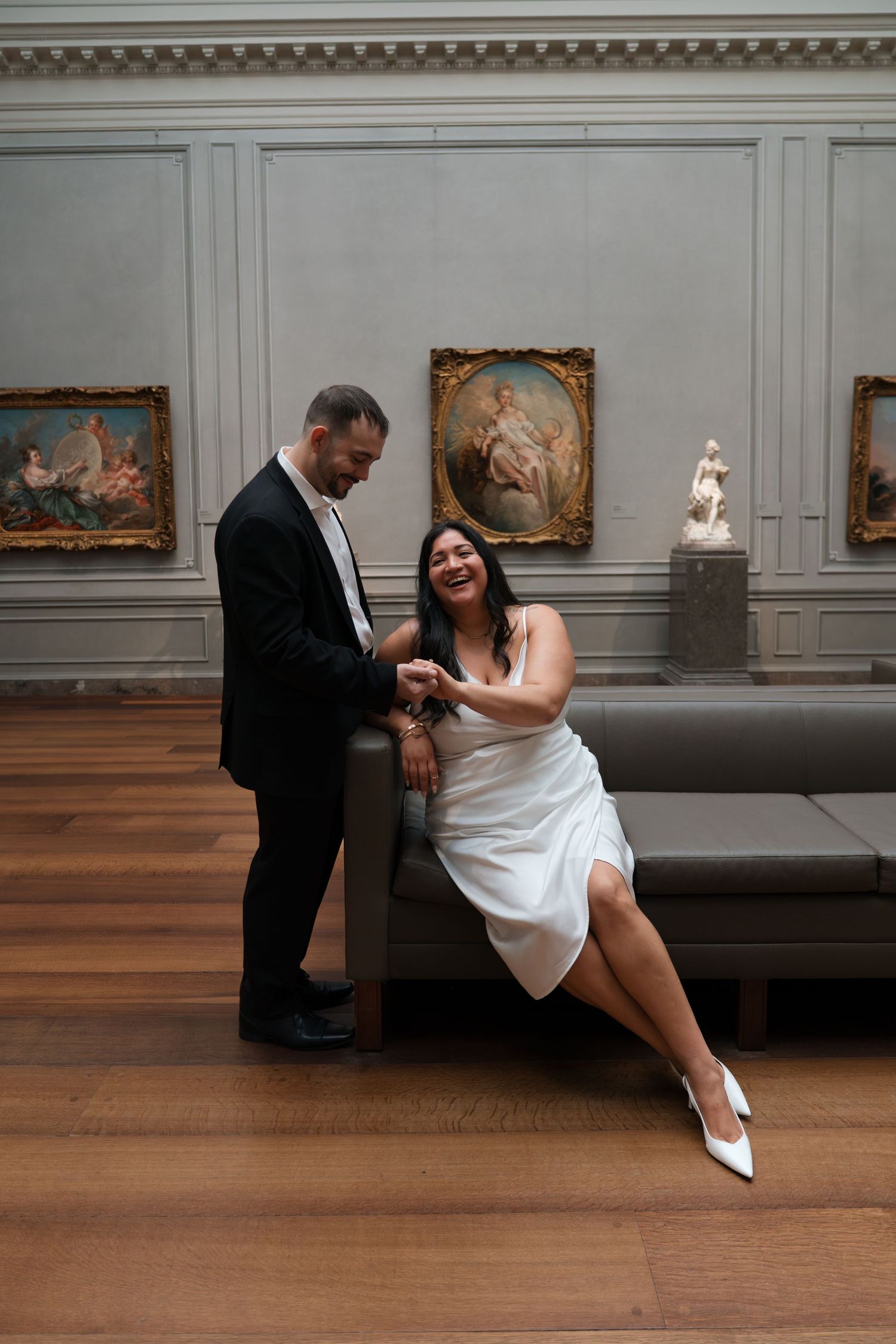 Couple in wedding attire laughing together in an elegant art museum gallery with classic paintings and hardwood floors.